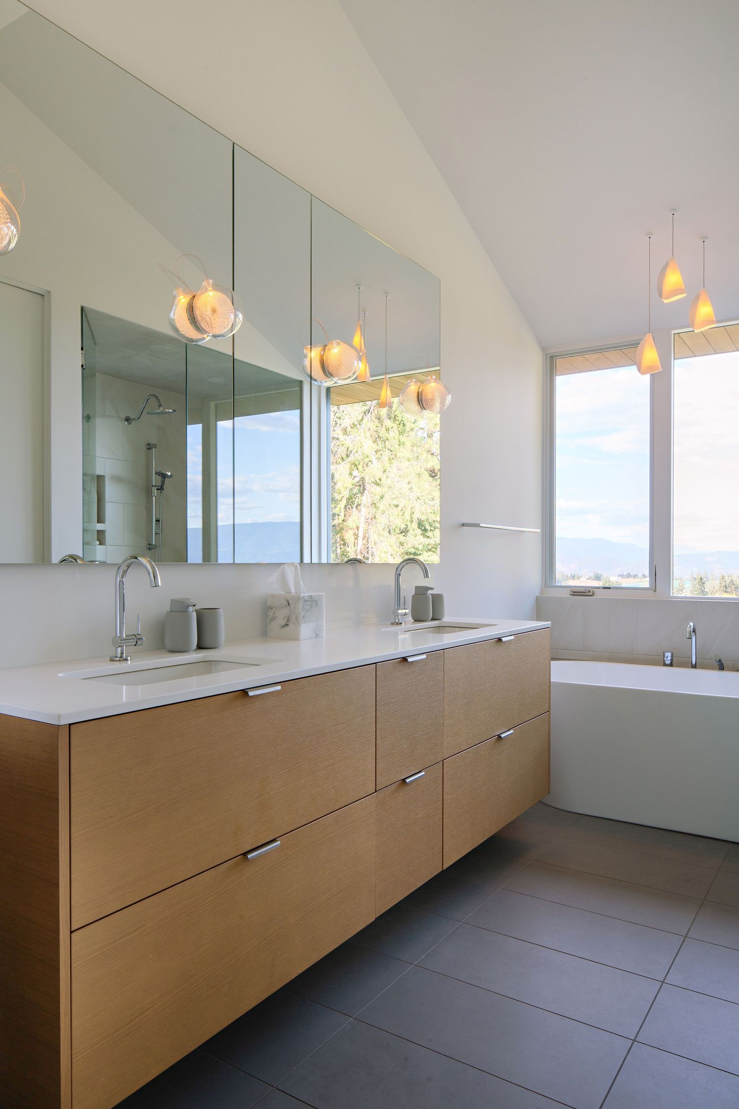 Modern bathroom interior with floating wood vanity, white countertop, pendant lights and freestanding tub near window.