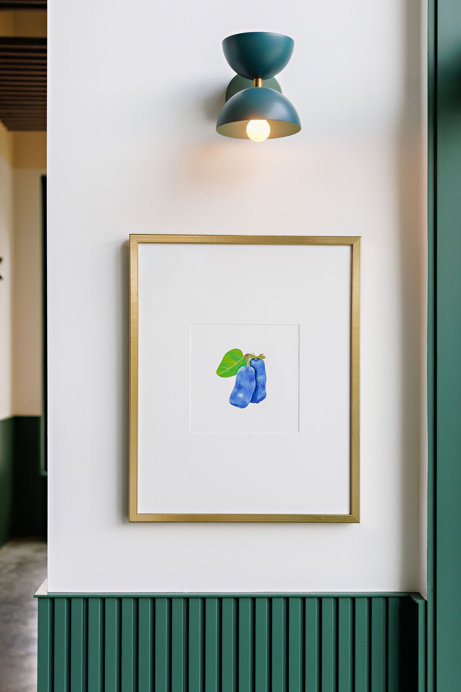 A minimalist art print of blue clothing hangs on a white wall beneath a teal ceiling light fixture mounted above.