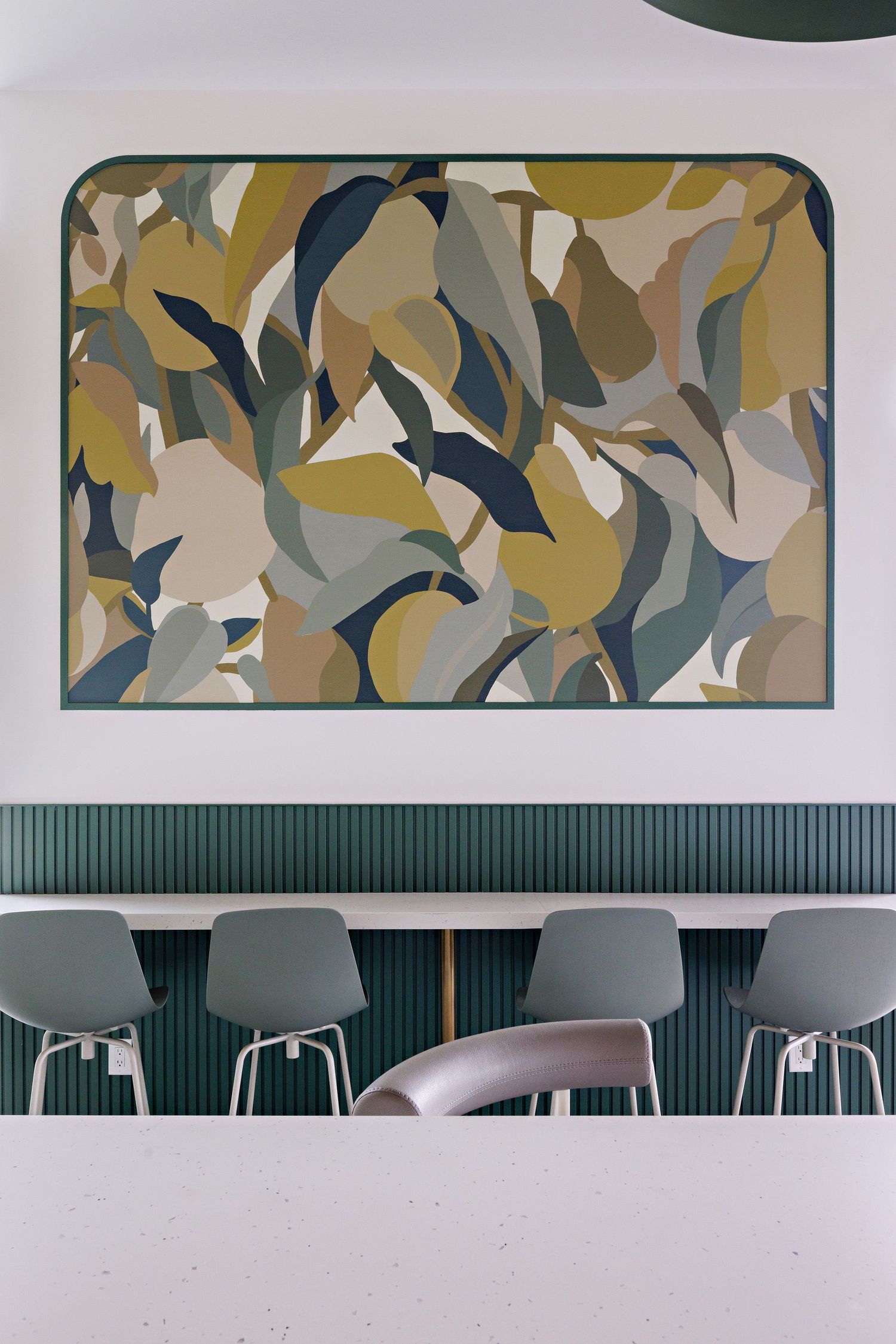 Modern abstract art piece with camouflage-like patterns in earthy tones mounted above green paneled wall and gray chairs.