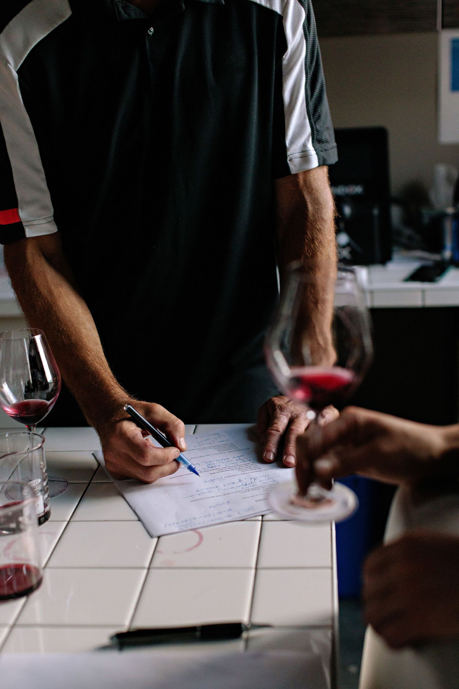 A person signs paperwork at a table with wine glasses while another person's hands are visible in the frame.