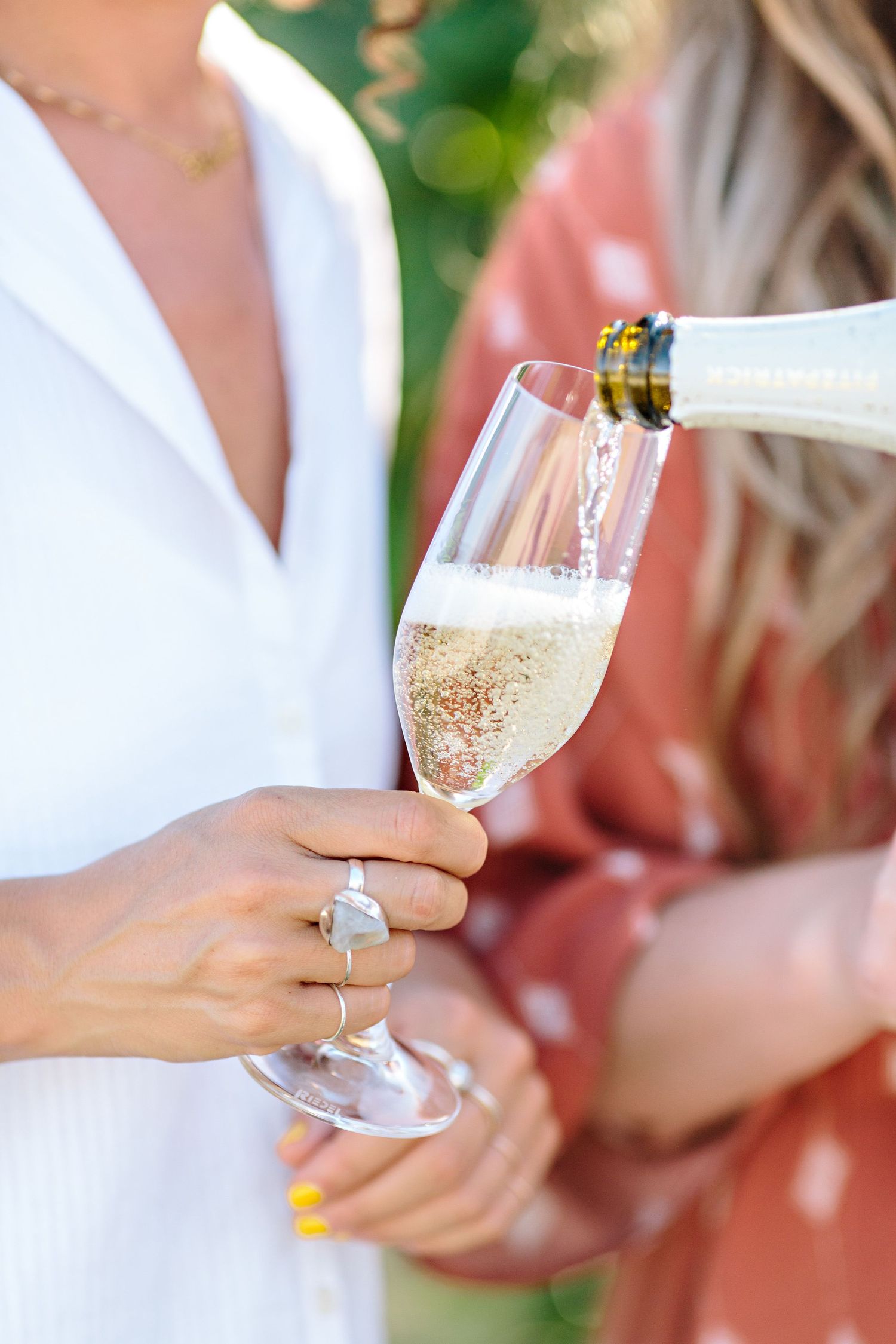 Champagne being poured into an elegant flute glass held by hand wearing yellow nail polish and a silver ring.