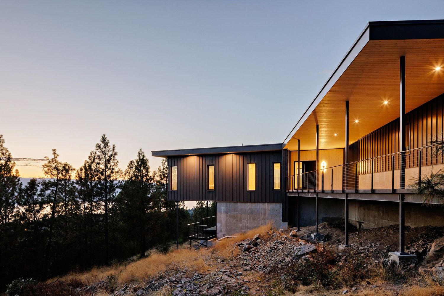 Modern hillside home with floor-to-ceiling windows glowing warmly at dusk against a mountainous backdrop.