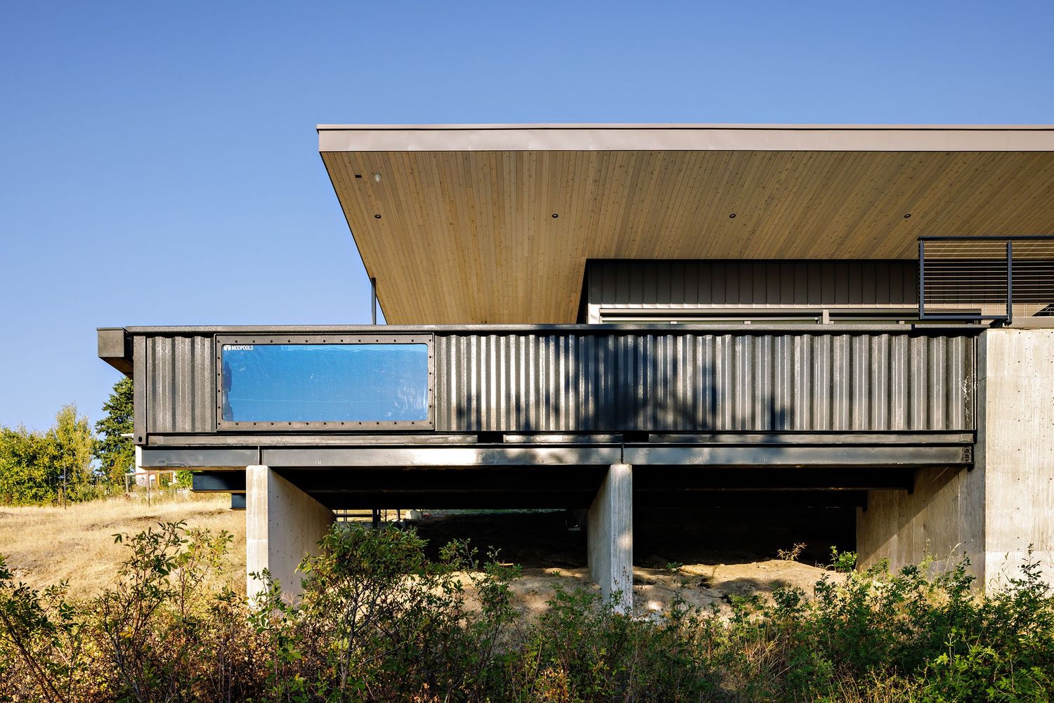 Modern hillside house with wooden overhanging deck supported by concrete pillars and glass railings against blue sky.