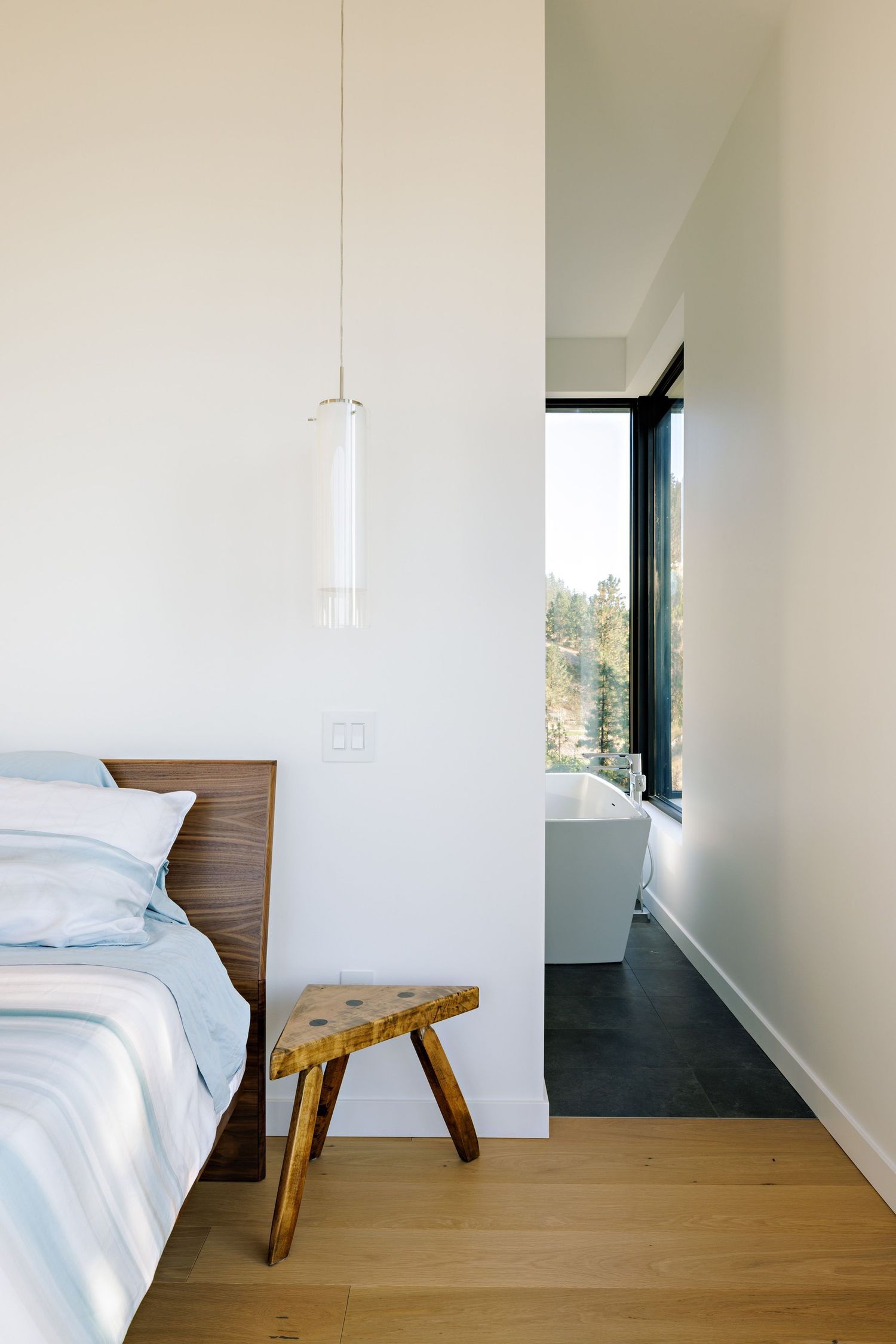 Minimalist bedroom with wooden bedframe and stool featuring a glimpse of a modern bathroom through a doorway.