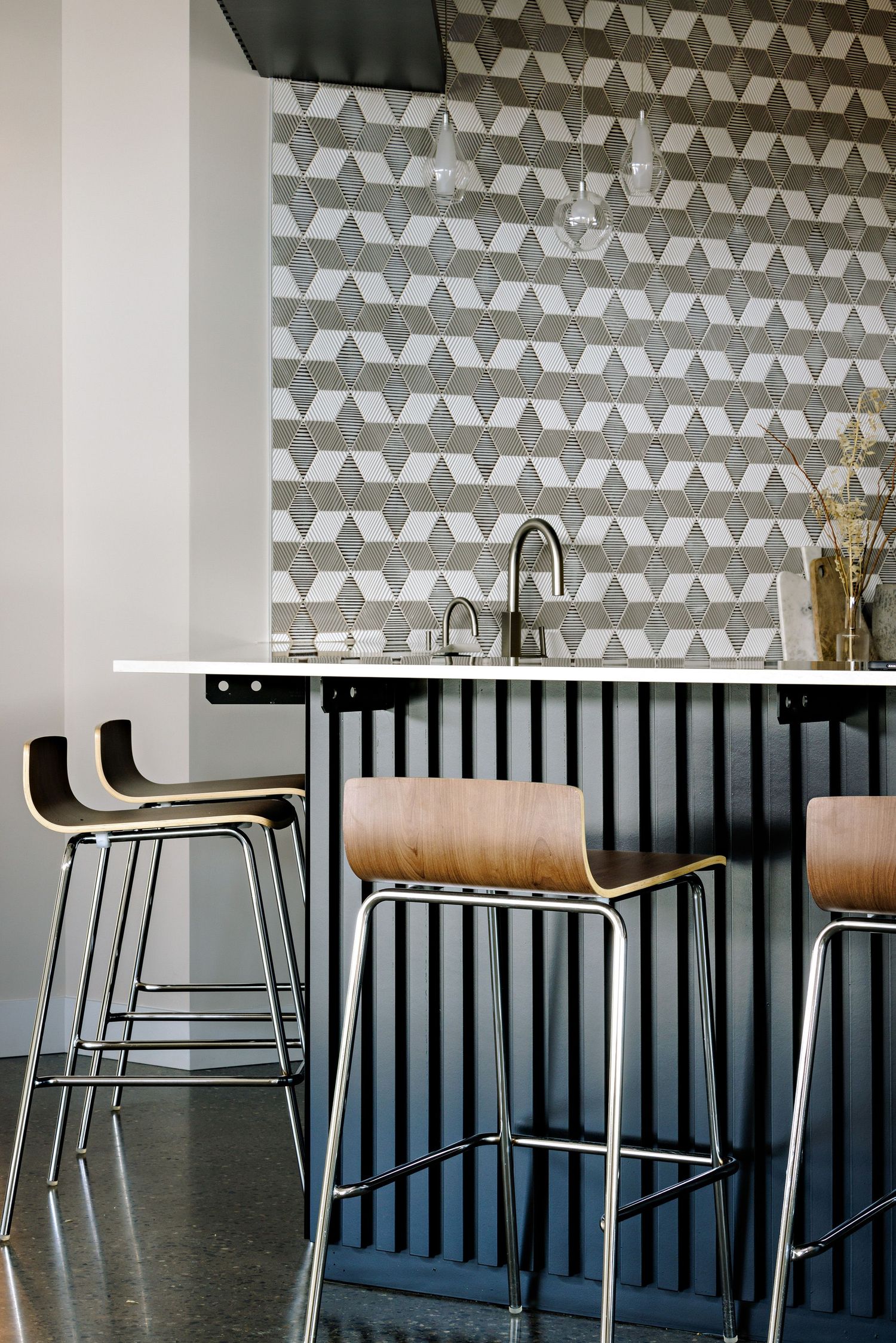 Modern bar stools with wooden seats line up against a dark counter beneath a geometric patterned accent wall.