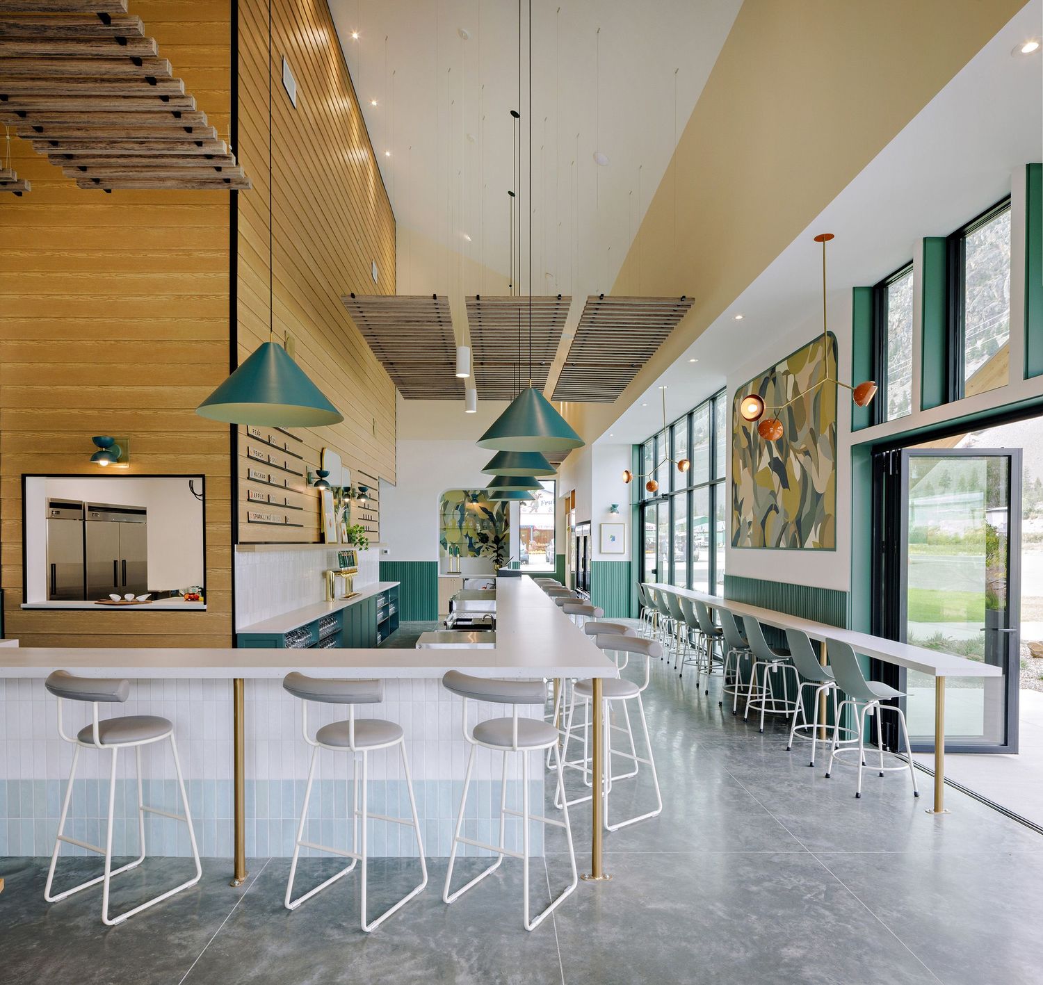Modern cafe interior with turquoise pendant lamps, white bar stools, and floor-to-ceiling windows looking out to green space.