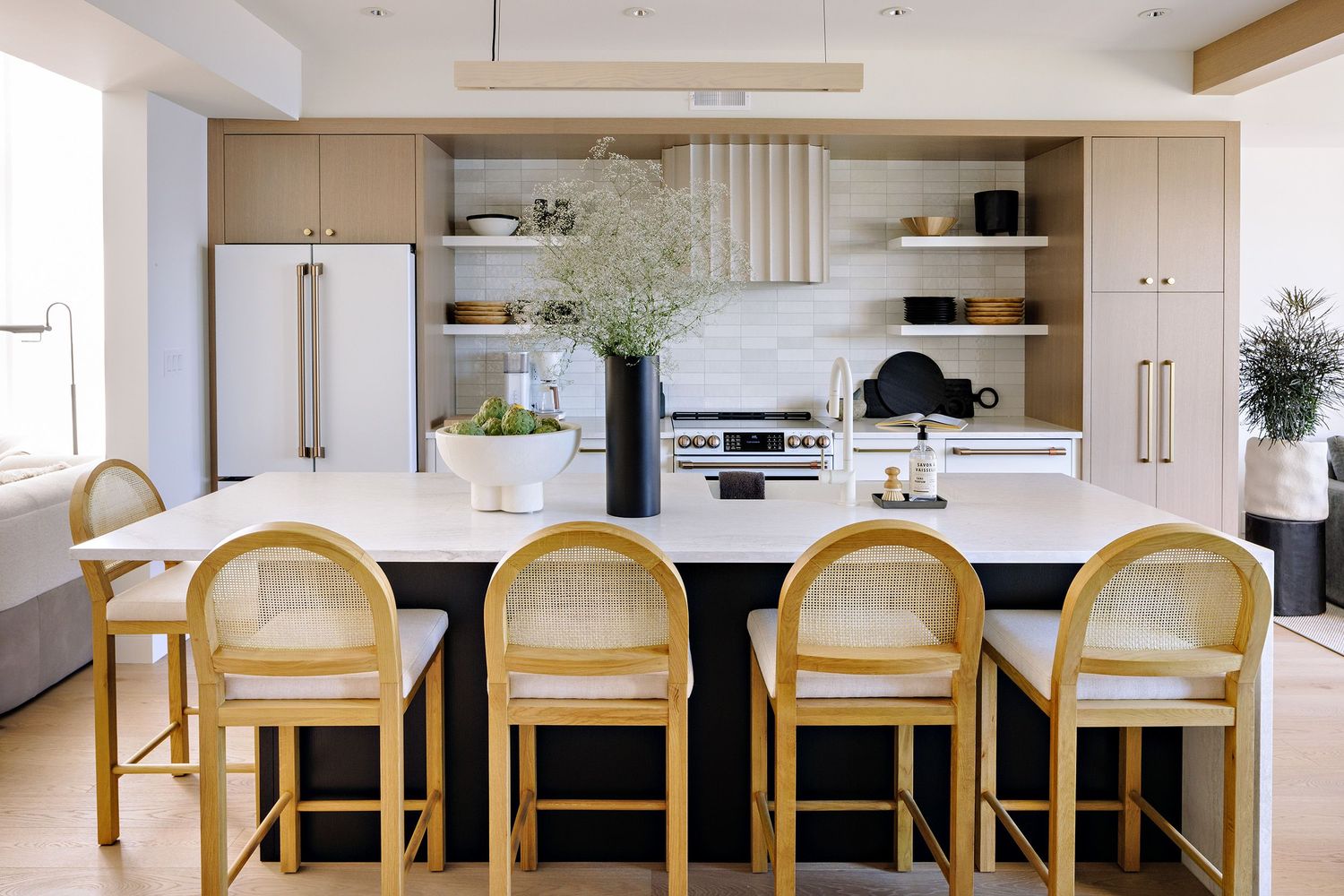Modern white kitchen with rattan bar stools and minimalist open shelving features sleek appliances and a large island.