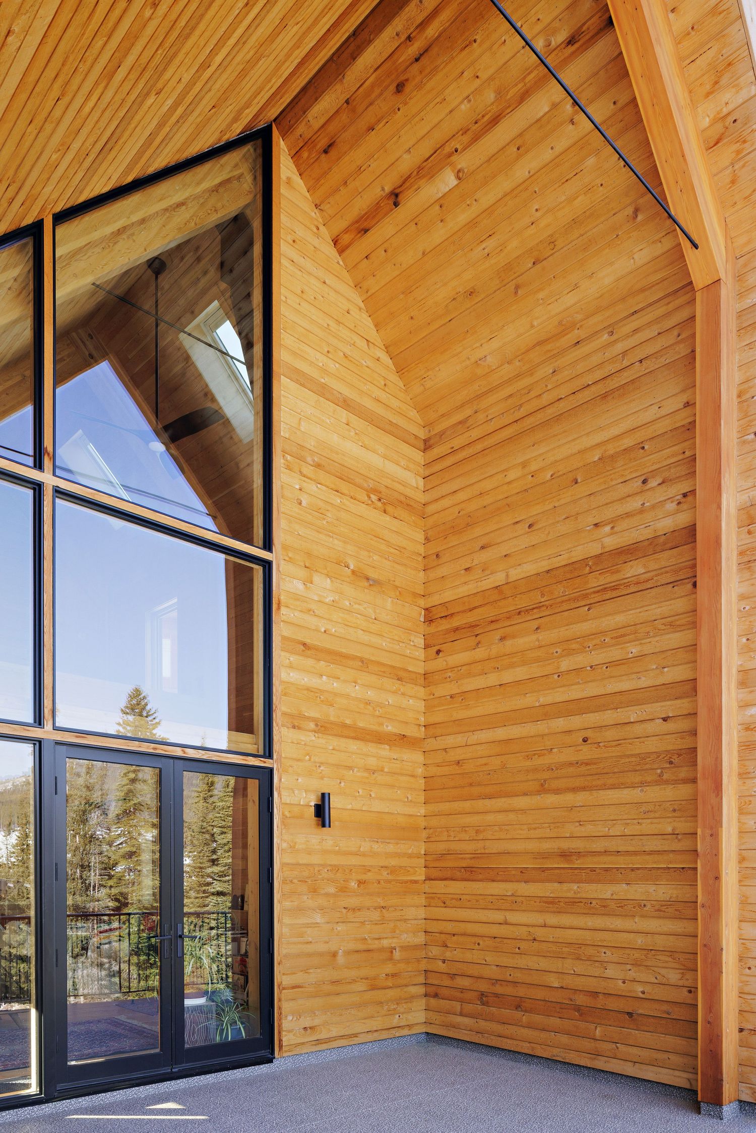 Modern cabin entrance features floor-to-ceiling windows and natural wood paneling against blue sky and forest views.