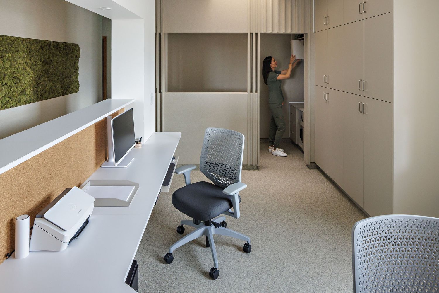 Modern office interior with white walls, grey carpet, and ergonomic furniture showing multiple workstations and seating areas.