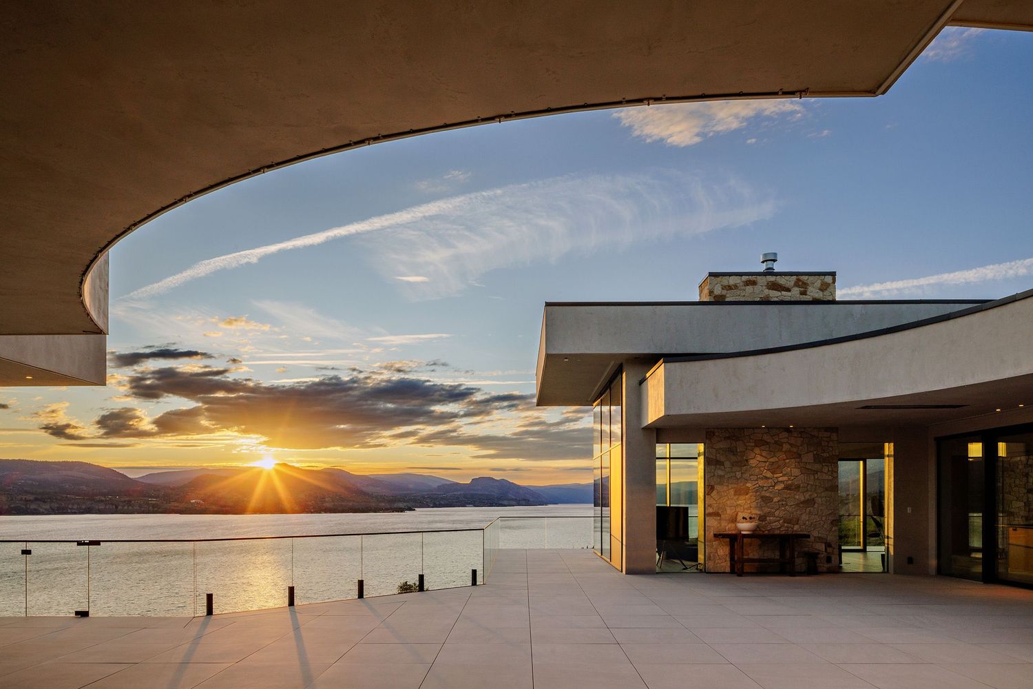 Modern architectural building with curved overhang framing a dramatic sunset over water and mountains.