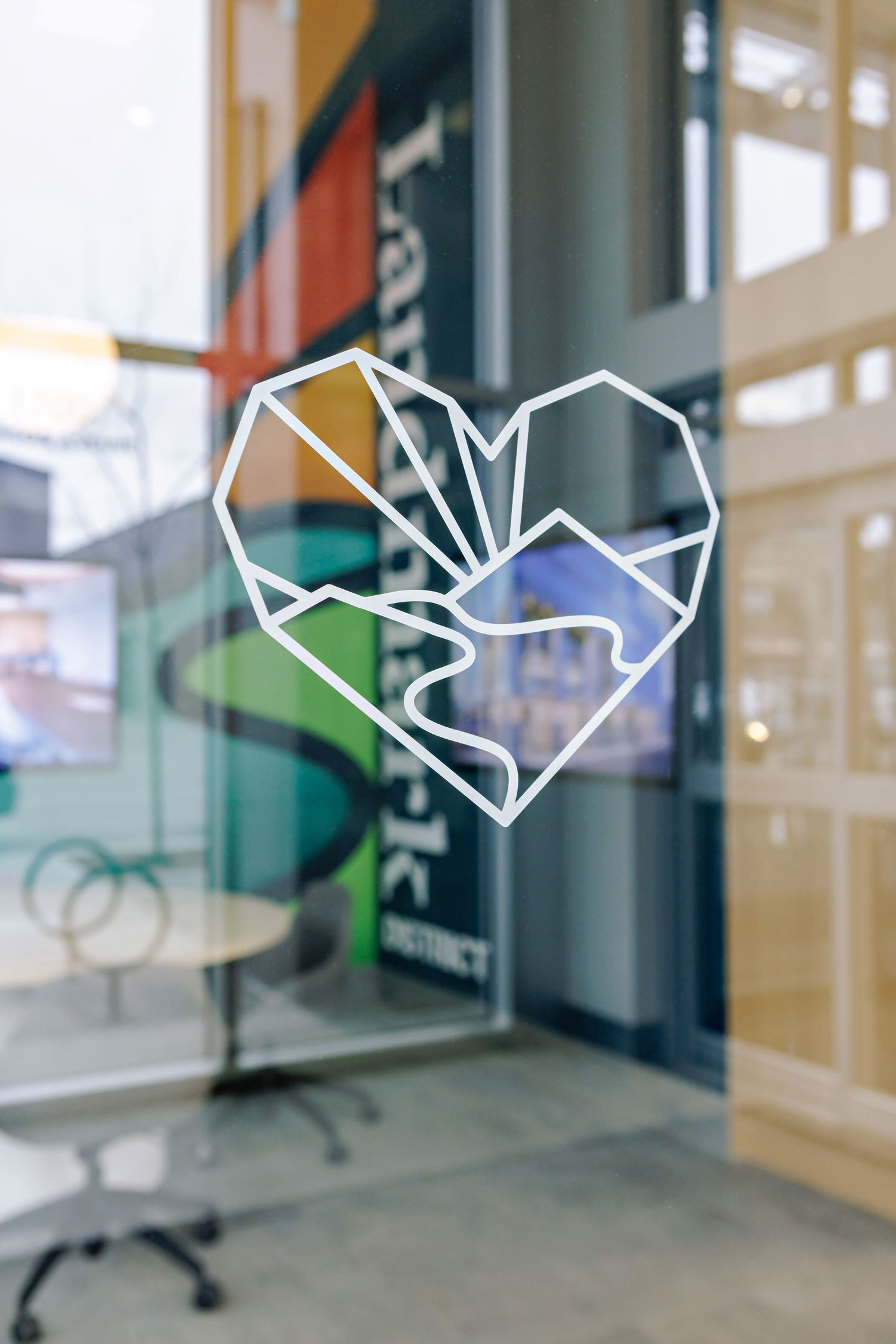 A geometric heart logo design in white lines on a glass window or door with blurred office interior background.