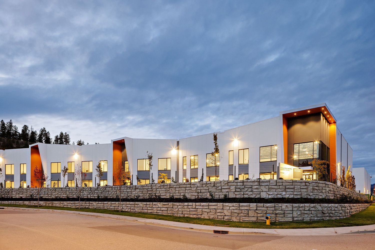 Modern commercial building with orange accents and stone retaining wall under dramatic cloudy sky at dusk.