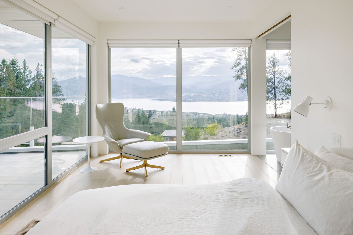 Modern minimalist bedroom with floor-to-ceiling windows offering stunning lake views, featuring a white wingback chair and bright decor.