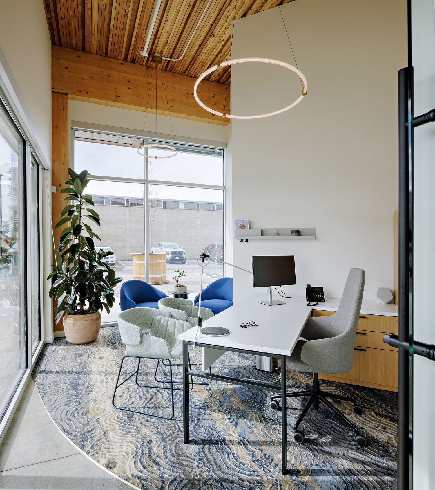 Modern office meeting room with white table, blue and gray chairs, wooden ceiling, and circular pendant light on patterned area rug.