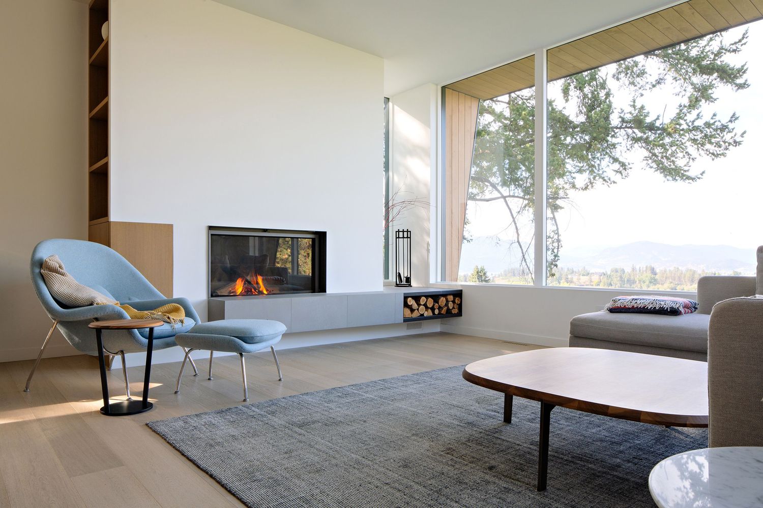 Contemporary living room with fireplace, gray armchair, and large windows offering scenic forest views.