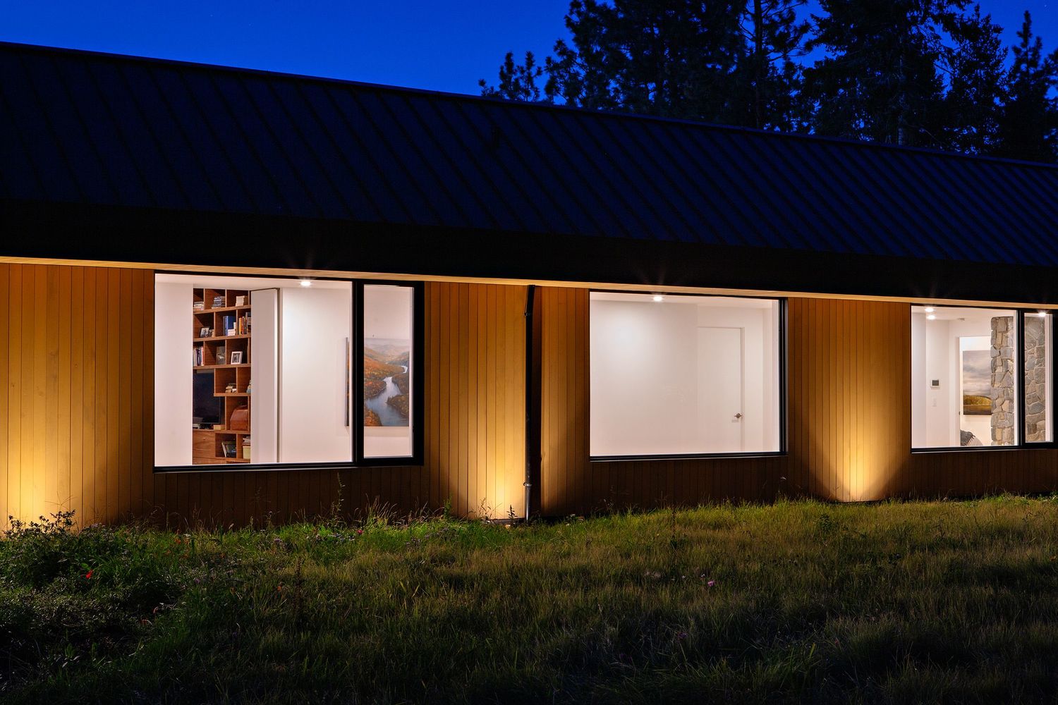 Modern ranch house exterior with warm uplighting and white windows glowing against wood siding at night.