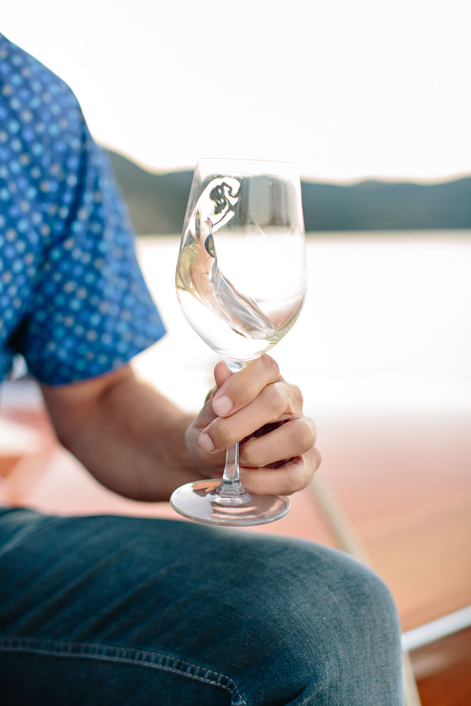 Person in blue shirt and jeans holding an empty wine glass by a waterfront view.
