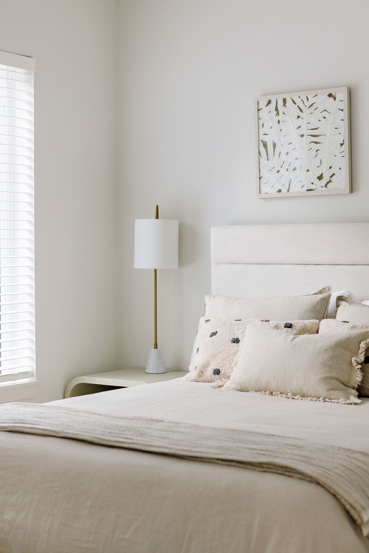 A serene neutral bedroom with white bedding, gold floor lamp, and minimalist botanical wall art above the headboard.