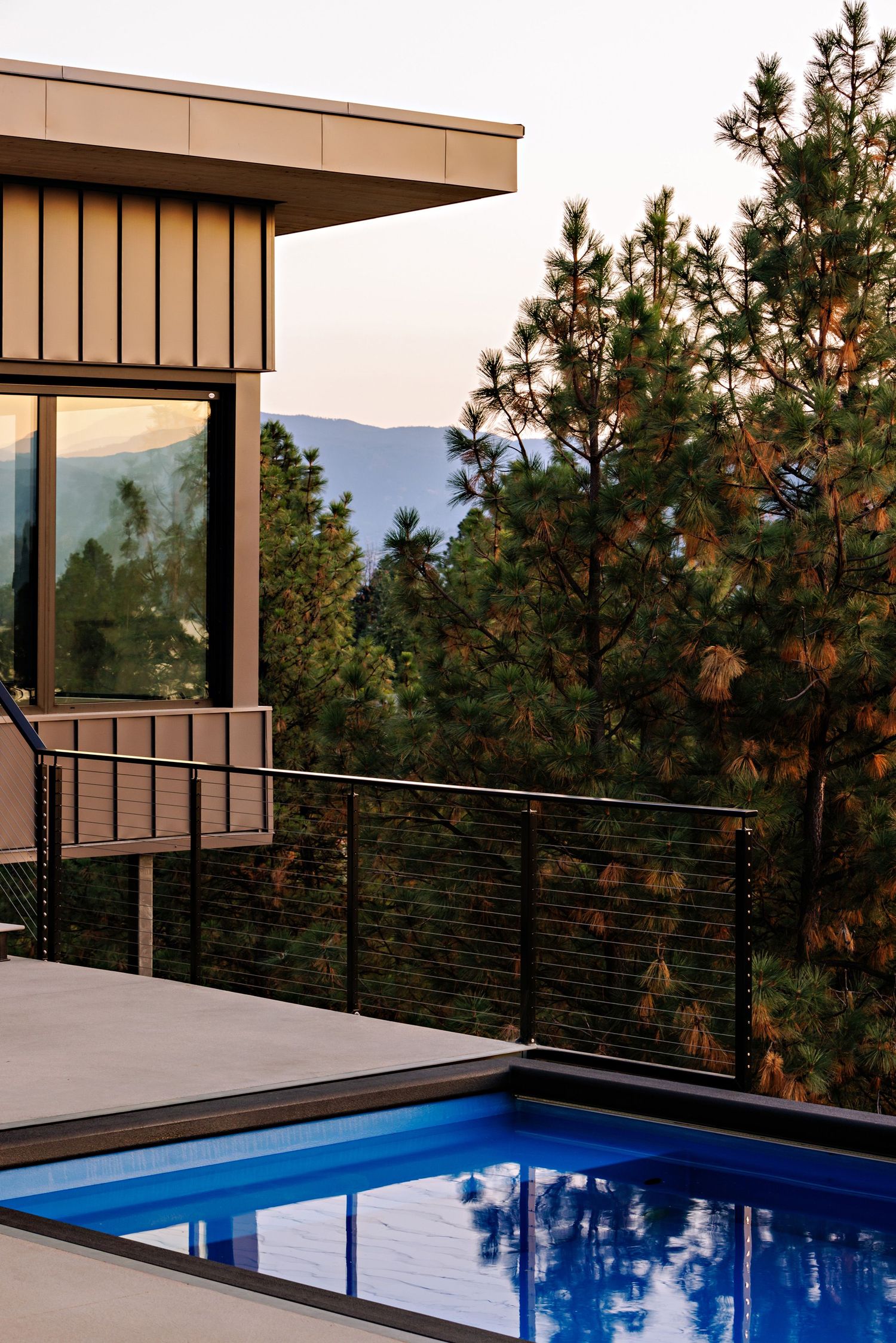 Modern luxury home with glass windows and stone facade features infinity pool overlooking mountain views at sunset.