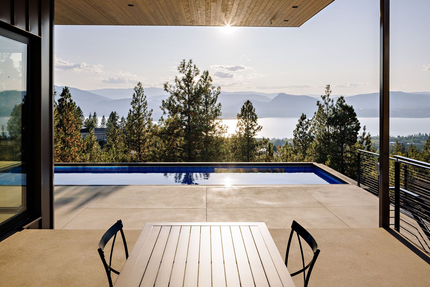 Luxury deck with infinity pool offers stunning mountain and lake views framed by tall pine trees and wood-paneled ceiling.