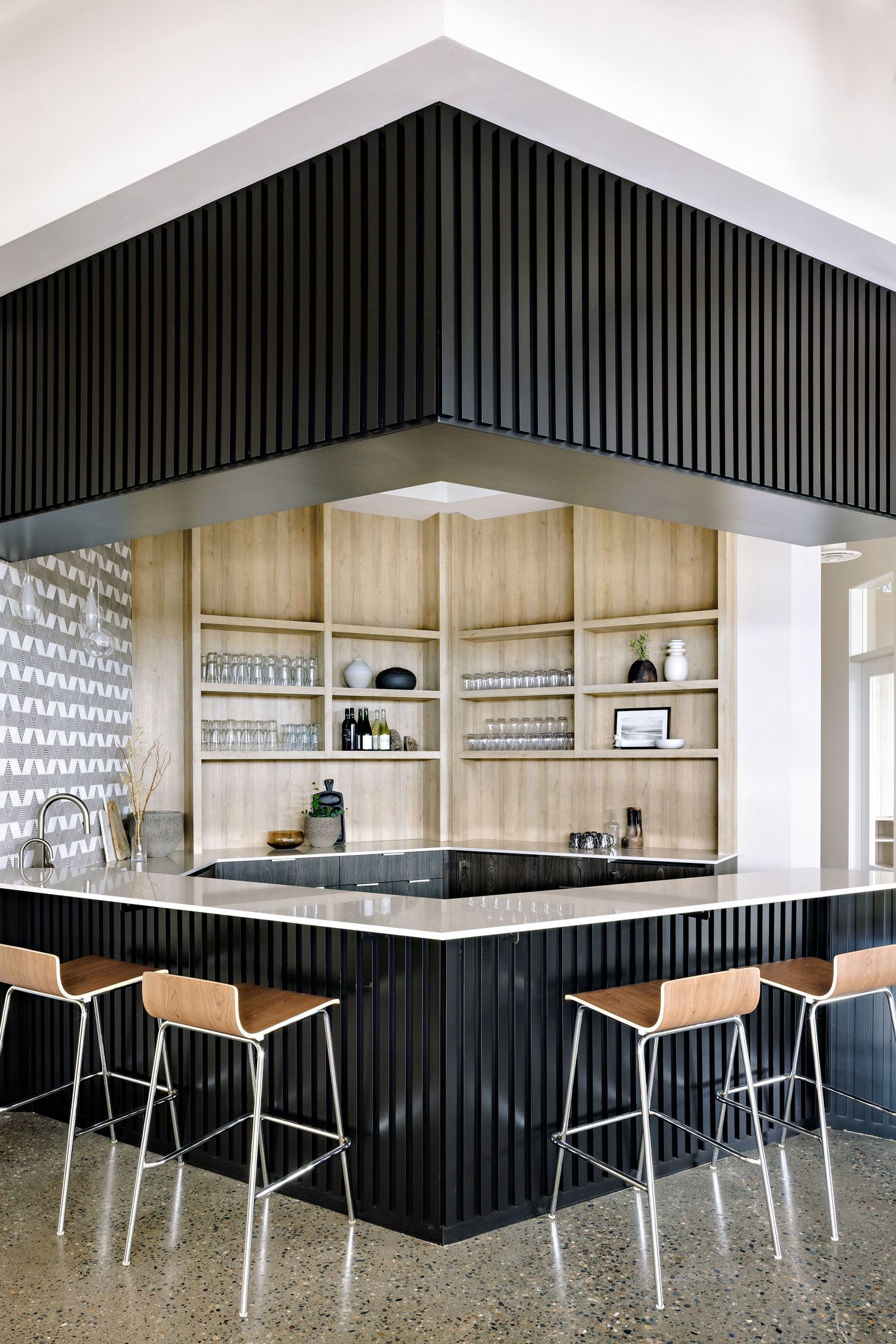 Modern kitchen bar with black ribbed paneling, wooden barstools, and built-in shelving displaying glassware and dishes.