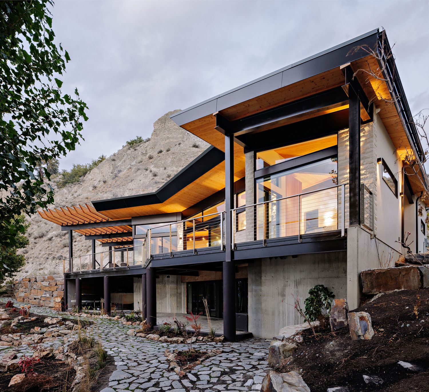 Modern hillside house with black exterior and large windows showcases dramatic angles against rocky terrain at dusk.