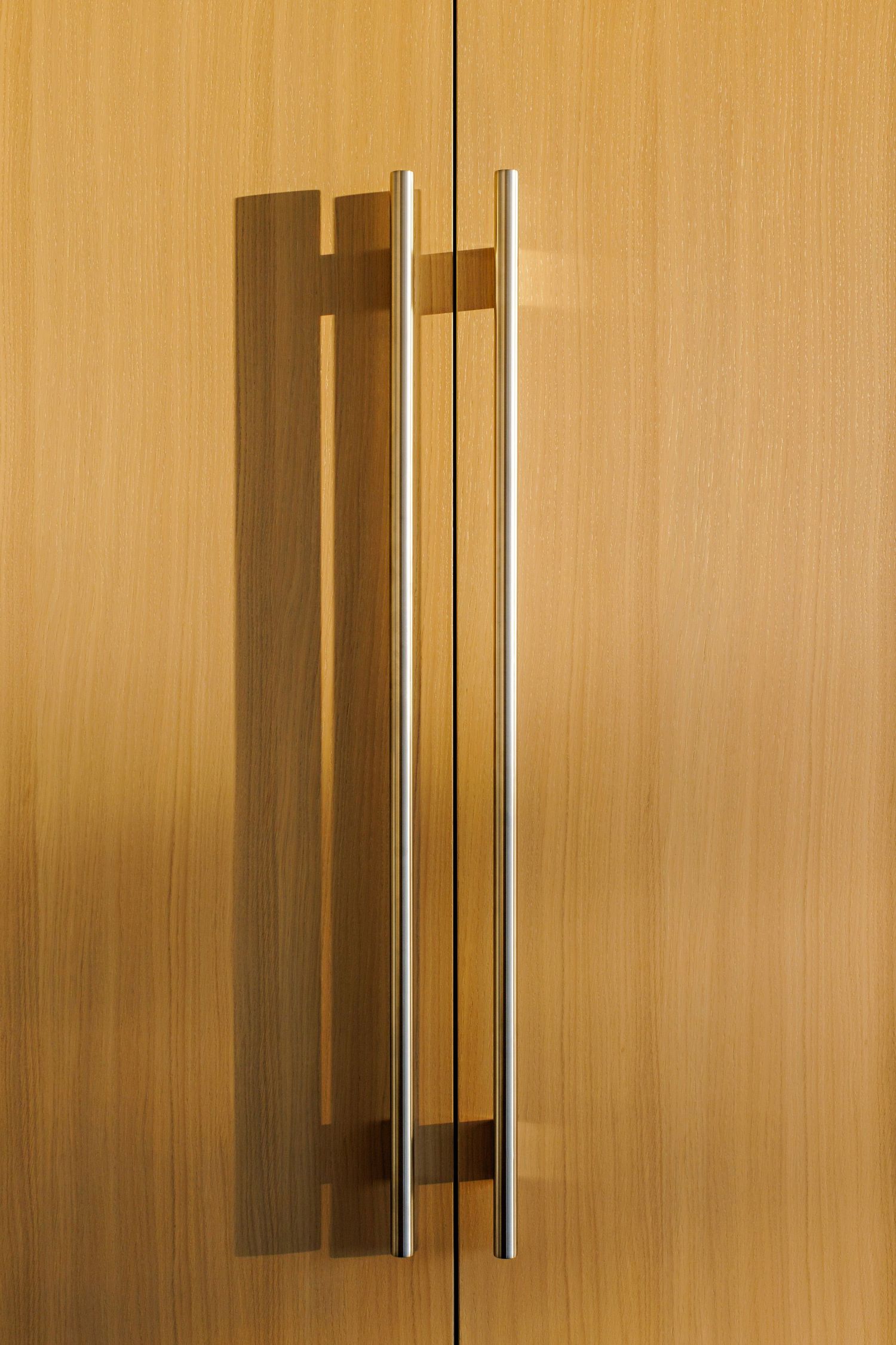 Modern stainless steel door handle with vertical bar design mounted on light wood cabinet surface.
