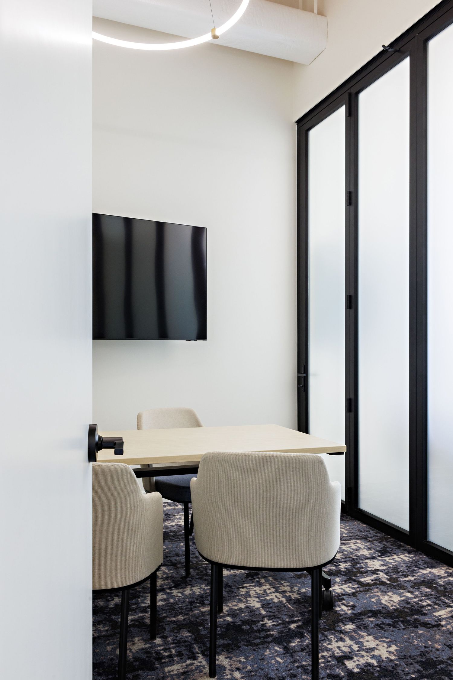 Modern office conference room with wall-mounted TV, beige chairs and patterned carpet against white walls and frosted glass.