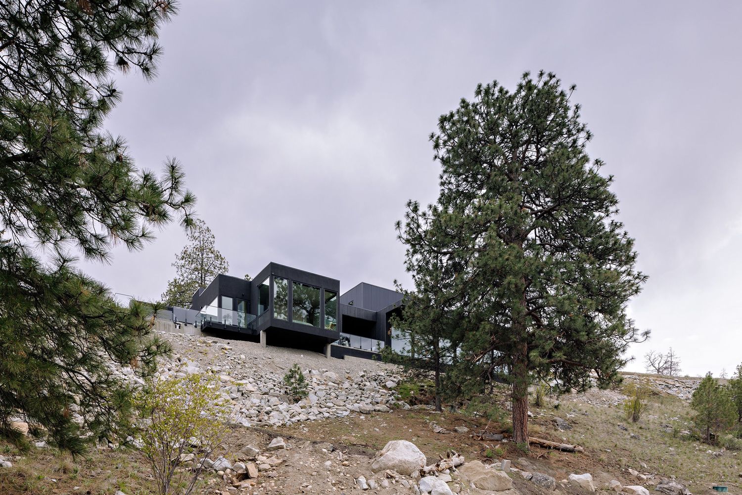 Modern black house perched dramatically on rocky hillside among pine trees with mountain views.