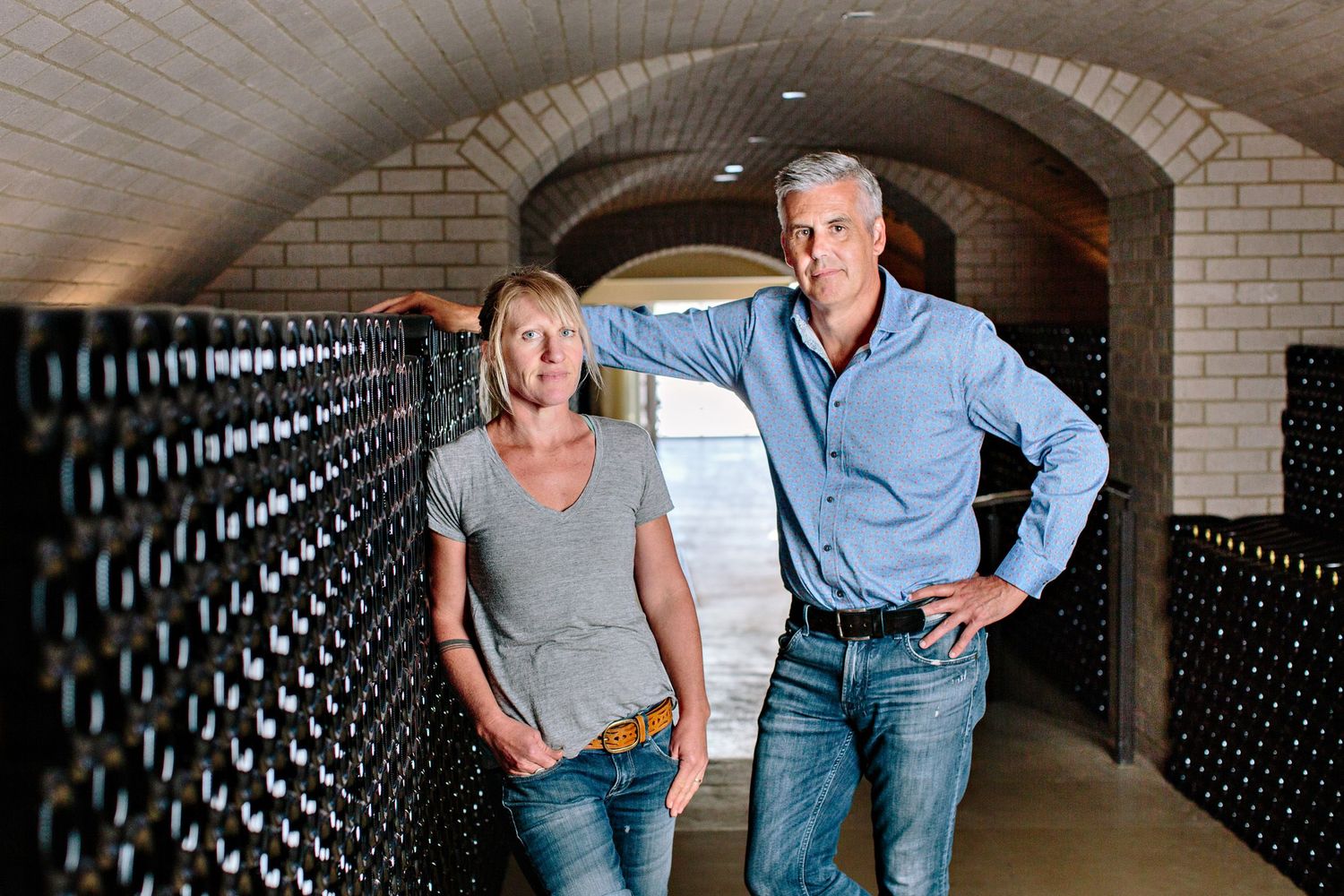 Two people standing in a wine cellar with curved brick ceiling and rows of bottles stored along the wall.