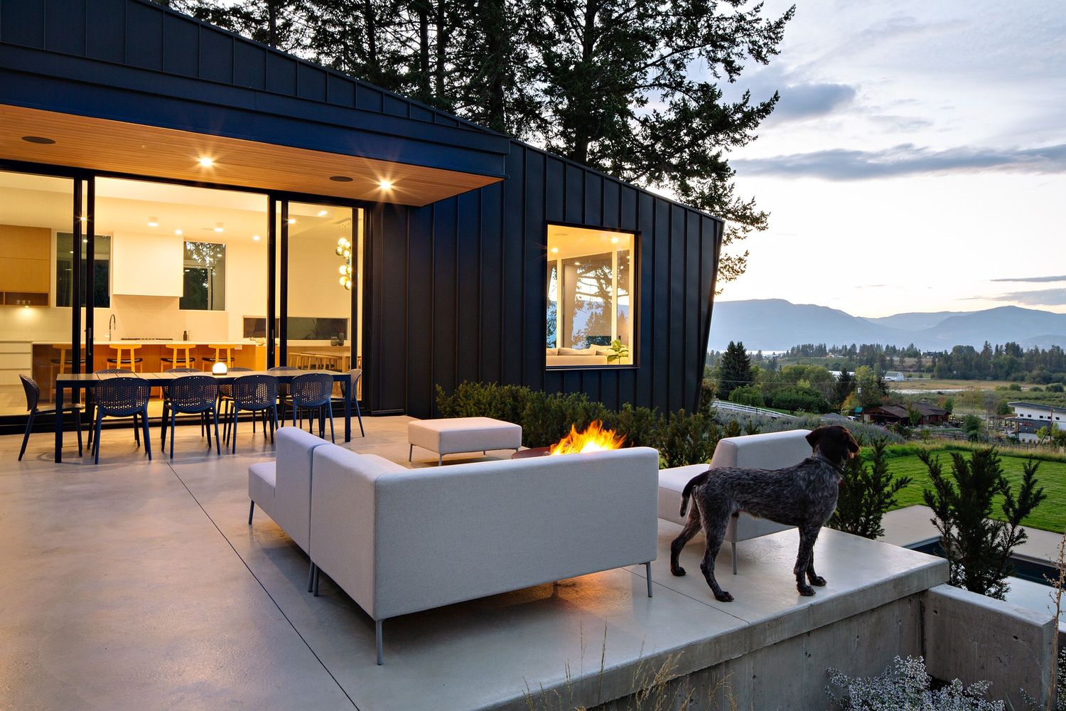 Contemporary house with large glass doors and outdoor lounge area featuring fire pit and panoramic valley views.