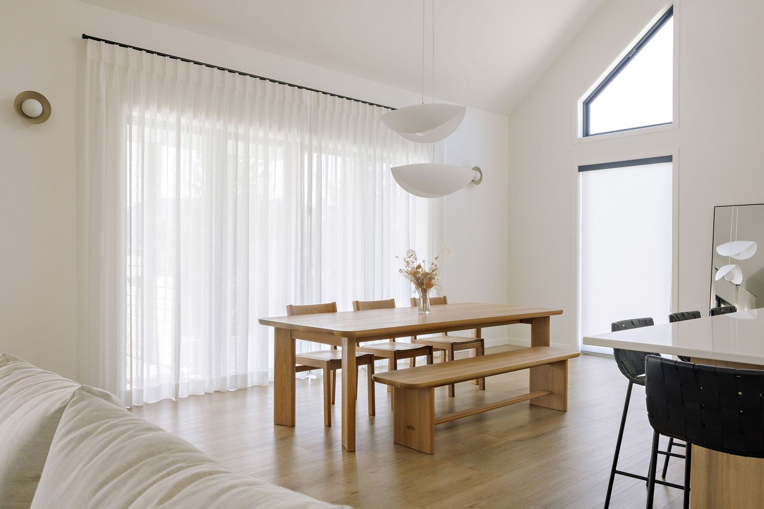 Modern minimalist dining area with wooden table and benches, white pendant light, and sheer curtains in bright interior space.