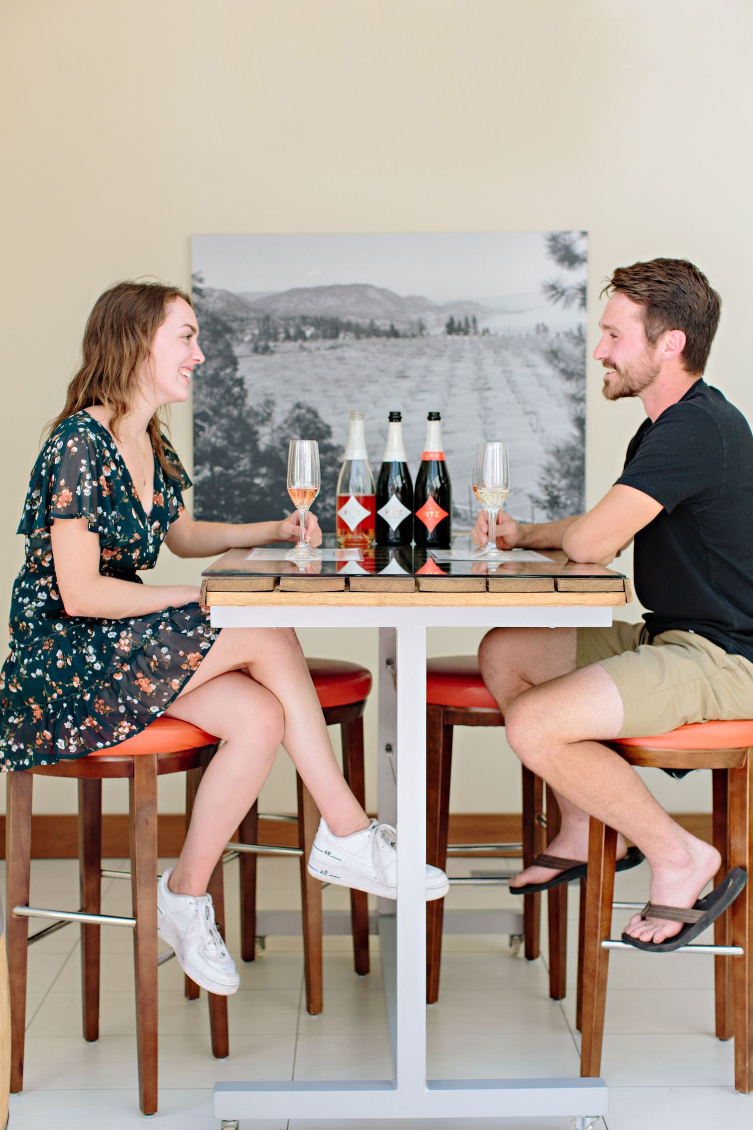 Two people enjoying drinks and conversation at a white bistro table with bottles and glassware.