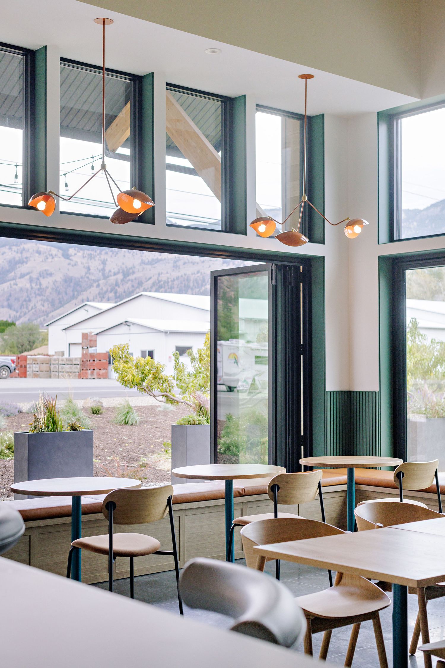 Modern cafe interior with floor-to-ceiling windows, pendant lights, and mountain views, featuring white tables and contemporary seating.