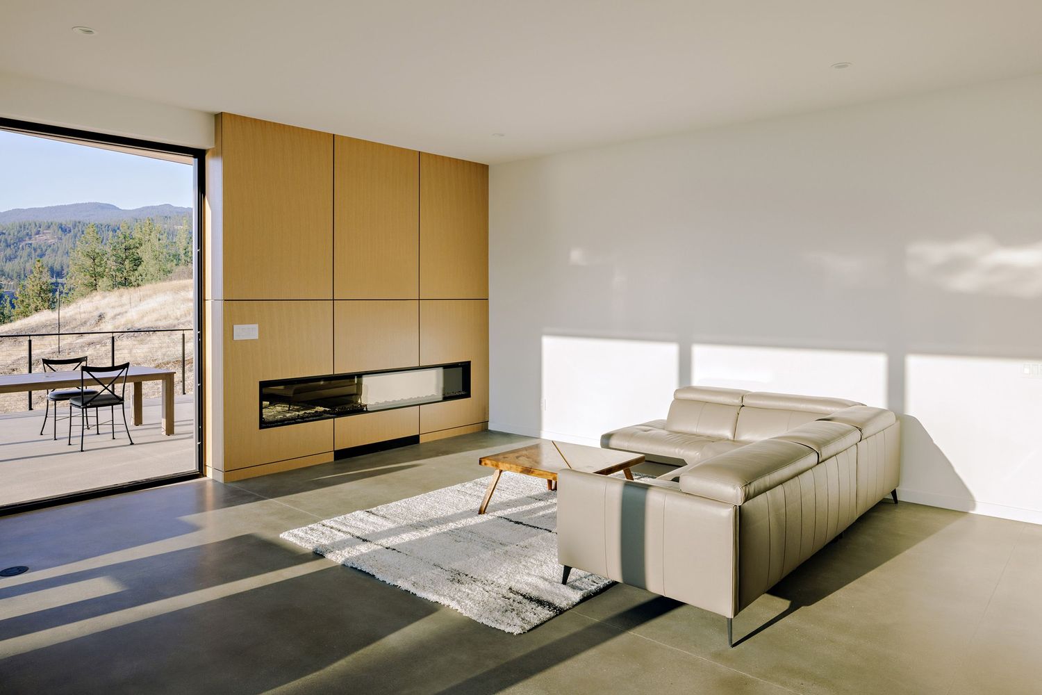 Modern minimalist living room with floor-to-ceiling windows, beige sectional sofa, and mountain views from balcony.
