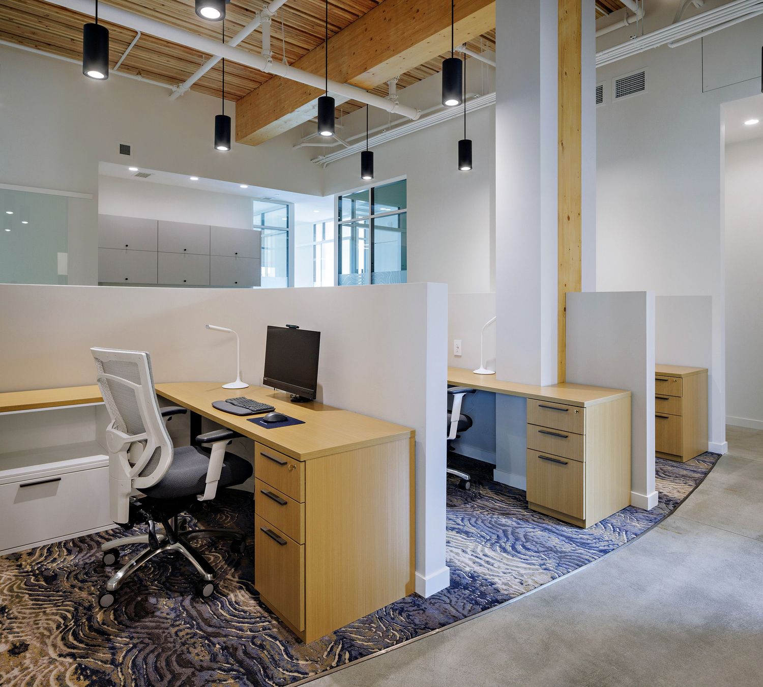 Modern office workspace with light wood desks, white chairs, and geometric patterned carpet in an open floor plan setting.