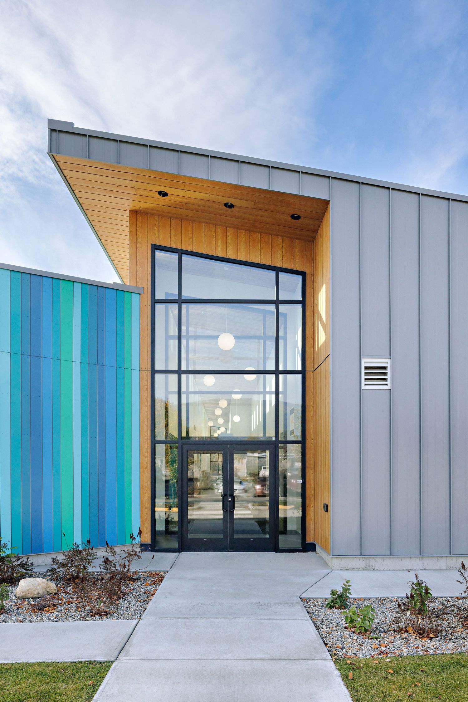 Modern building entrance with glass doors, gray metal siding, wood accent panel, and turquoise wall feature under blue sky.