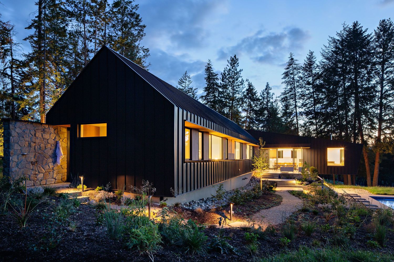Modern black wooden house with illuminated windows and pathway lights set against twilight forest backdrop in Pacific Northwest.
