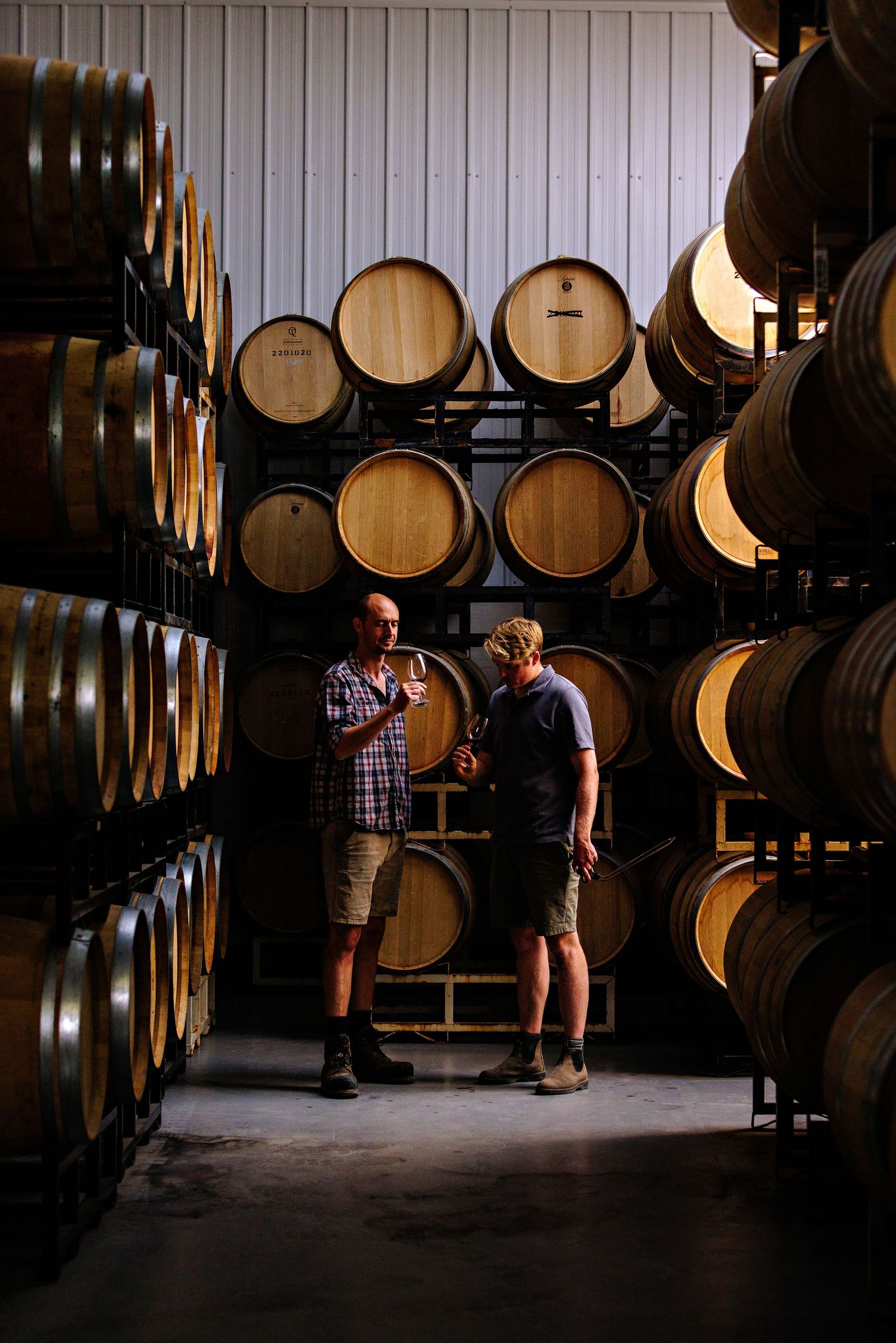 Two people sample wine while standing among stacked oak barrels in a dimly lit winery cellar.