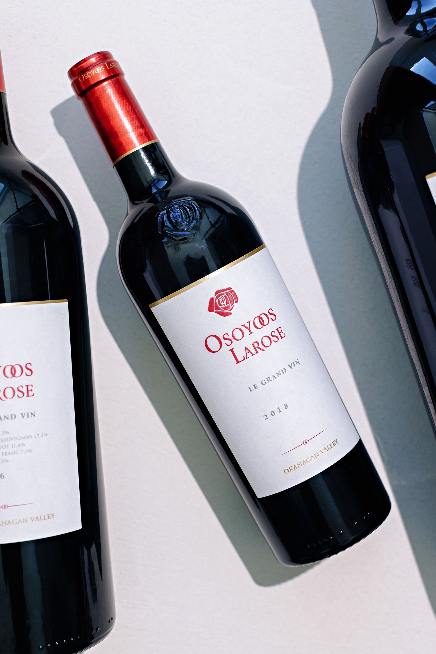 A bottle of Osoyoos Larose red wine casting shadows against a white surface alongside other wine bottles.
