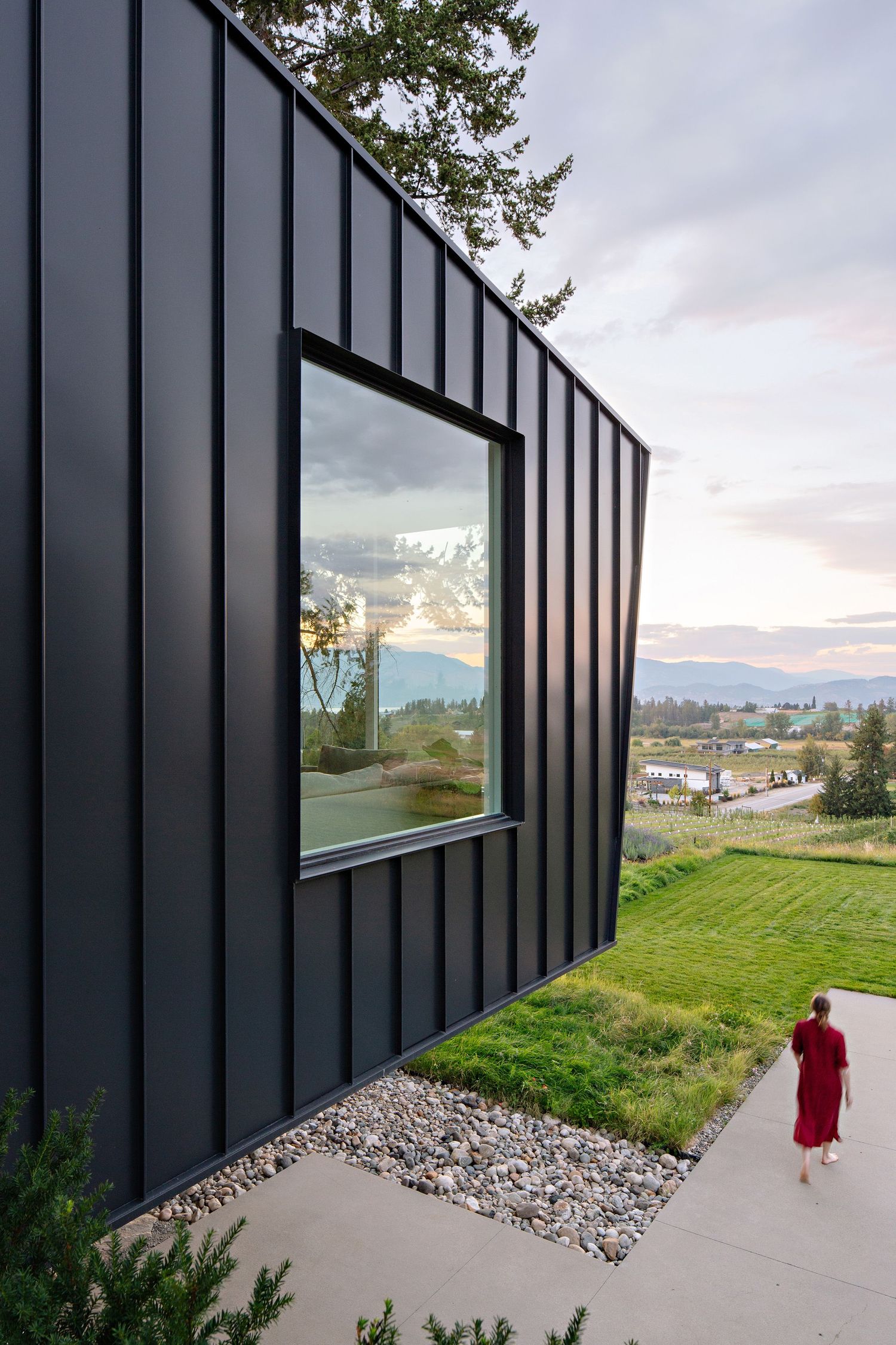 Modern black-clad house with large window reflects landscape views while someone walks along concrete path below.