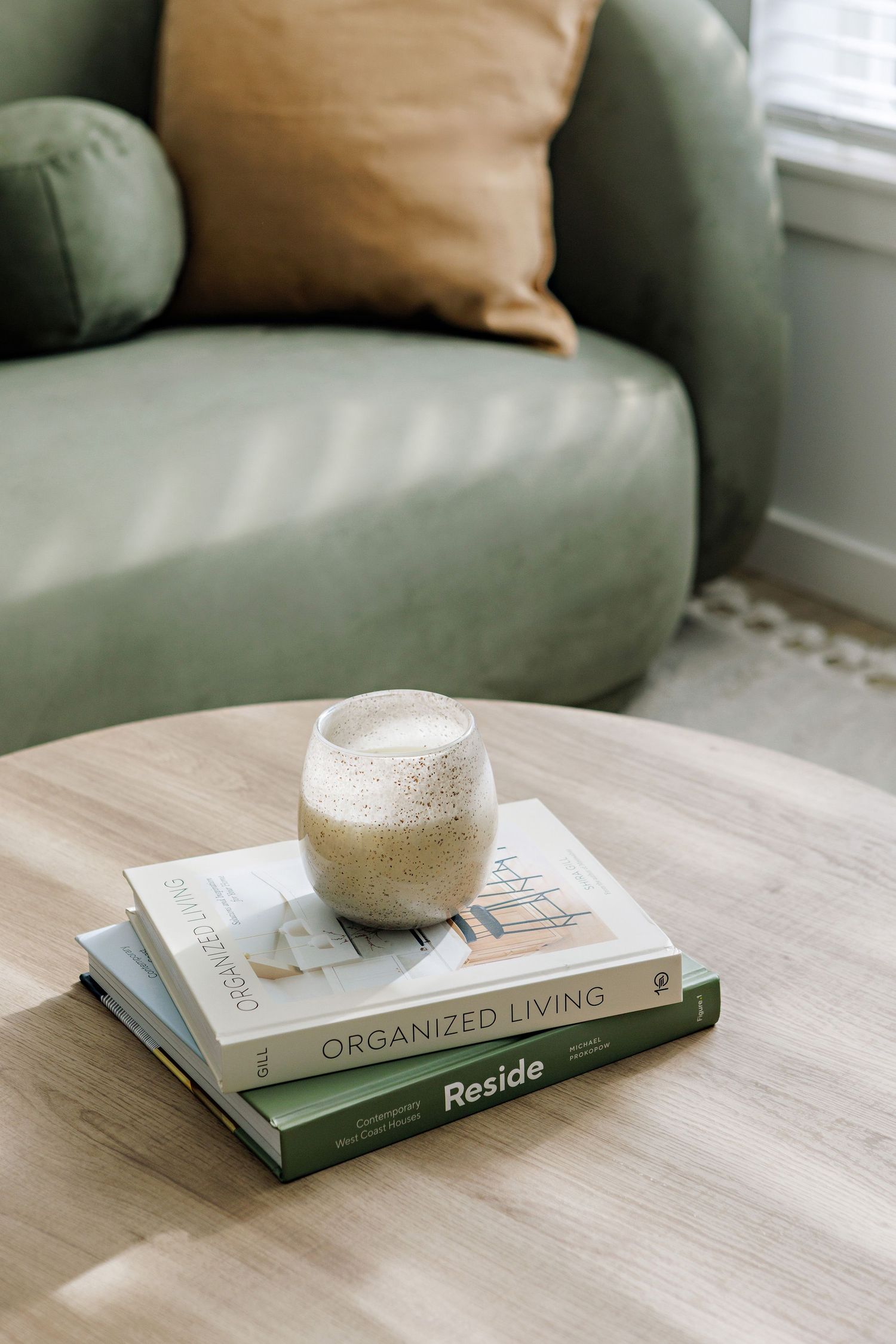 A creamy beverage sits in a glass atop two books about organized living on a wooden coffee table near a sage green couch.