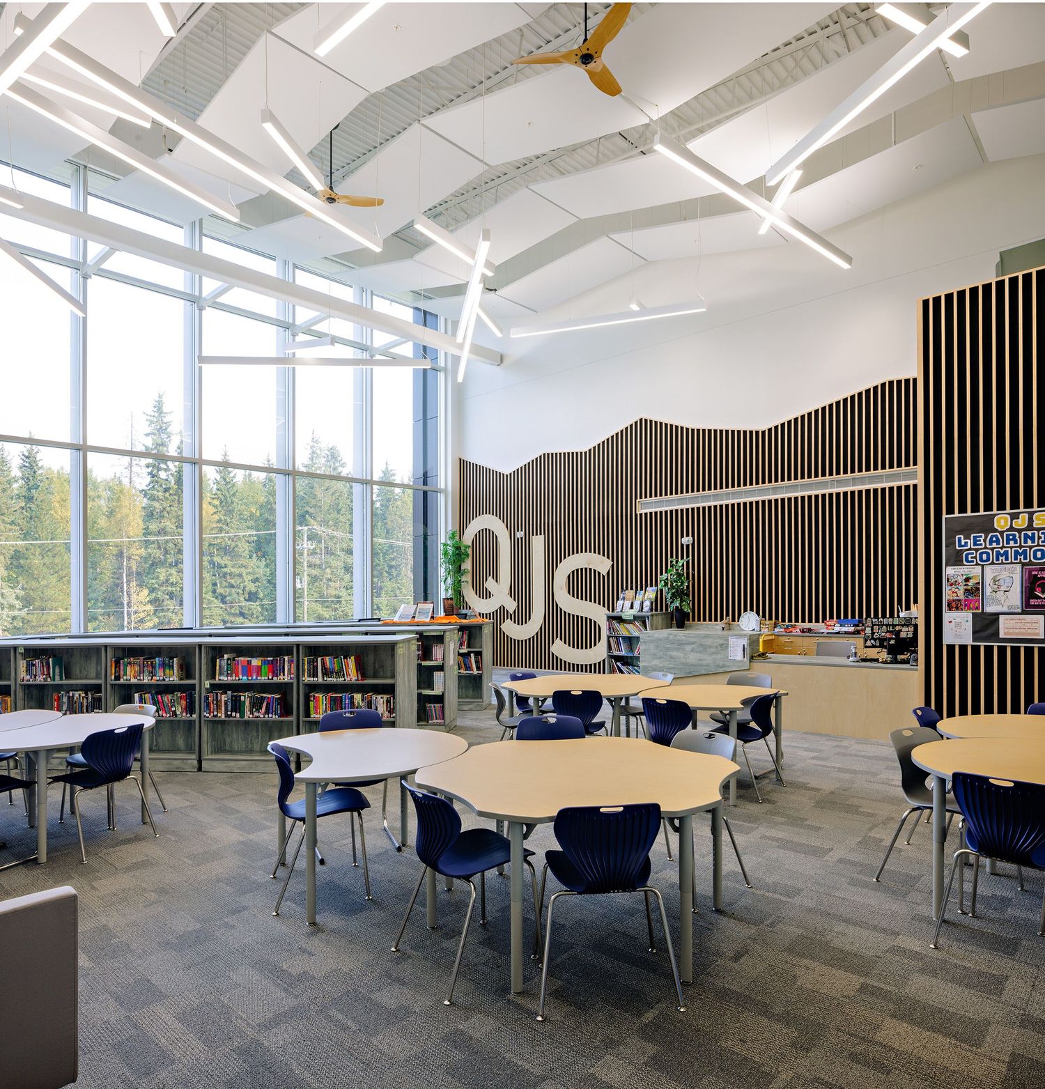 Modern school library interior with curved wooden wall feature, round tables, and large windows overlooking trees.