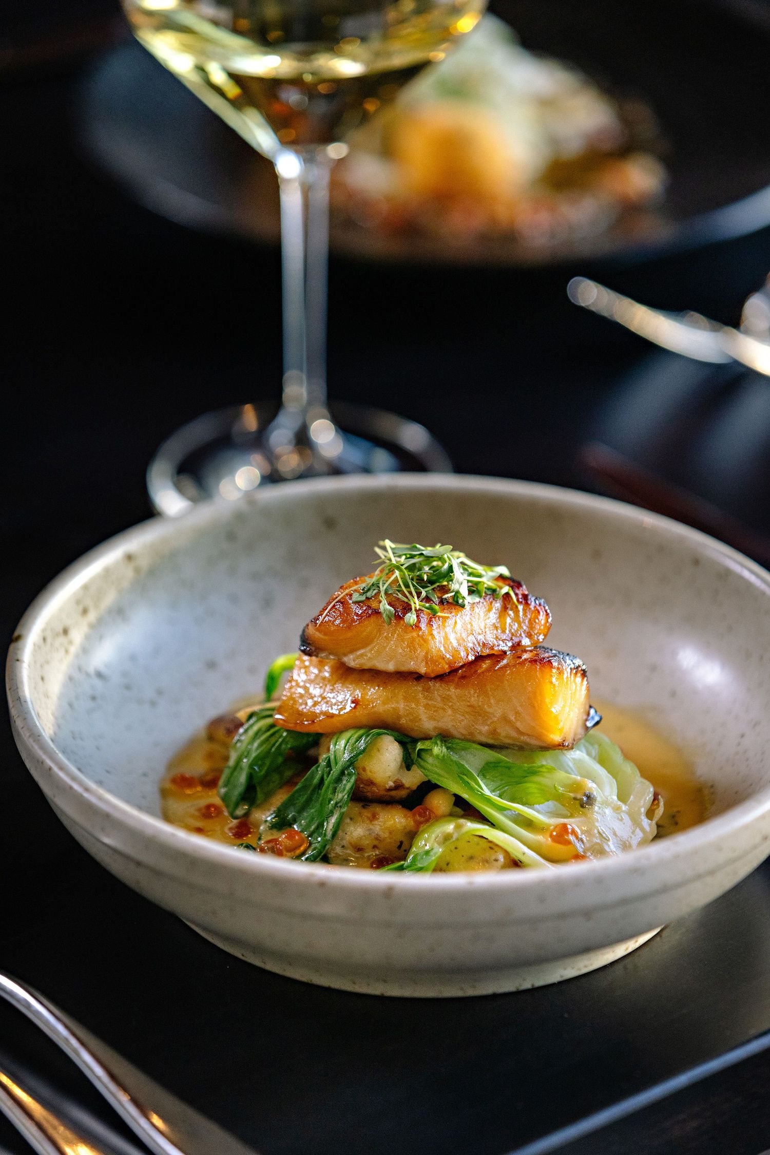Gourmet salmon dish with microgreens served in a ceramic bowl alongside a glass of white wine.