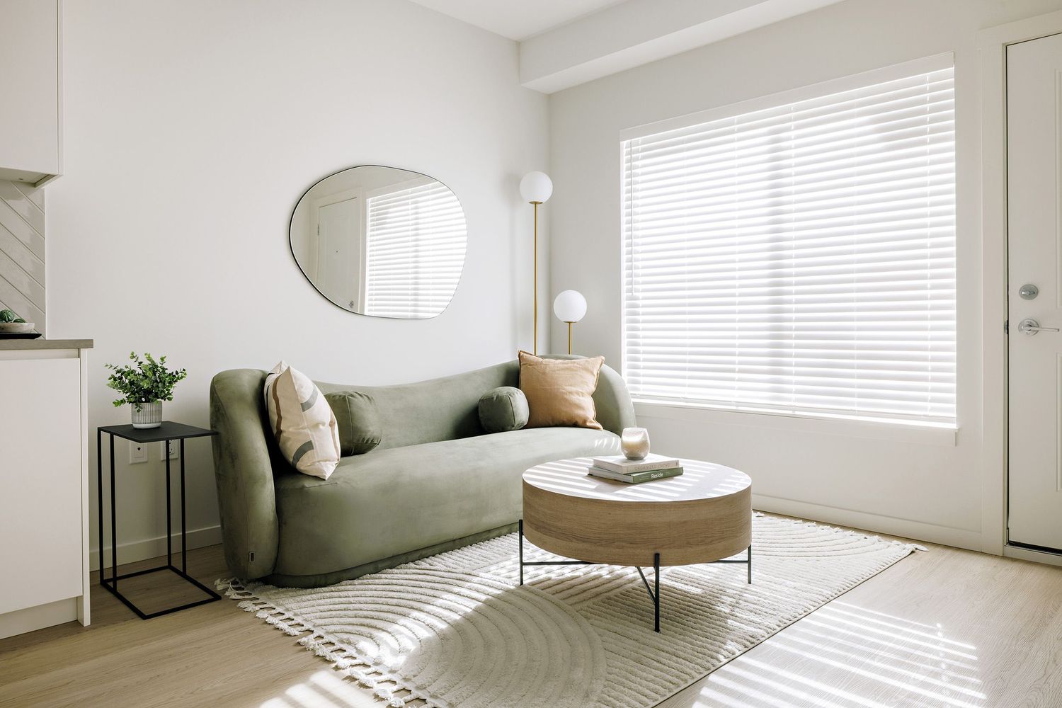 A minimalist living room with a sage green sofa, round coffee table, oval mirror, and natural light streaming through blinds.