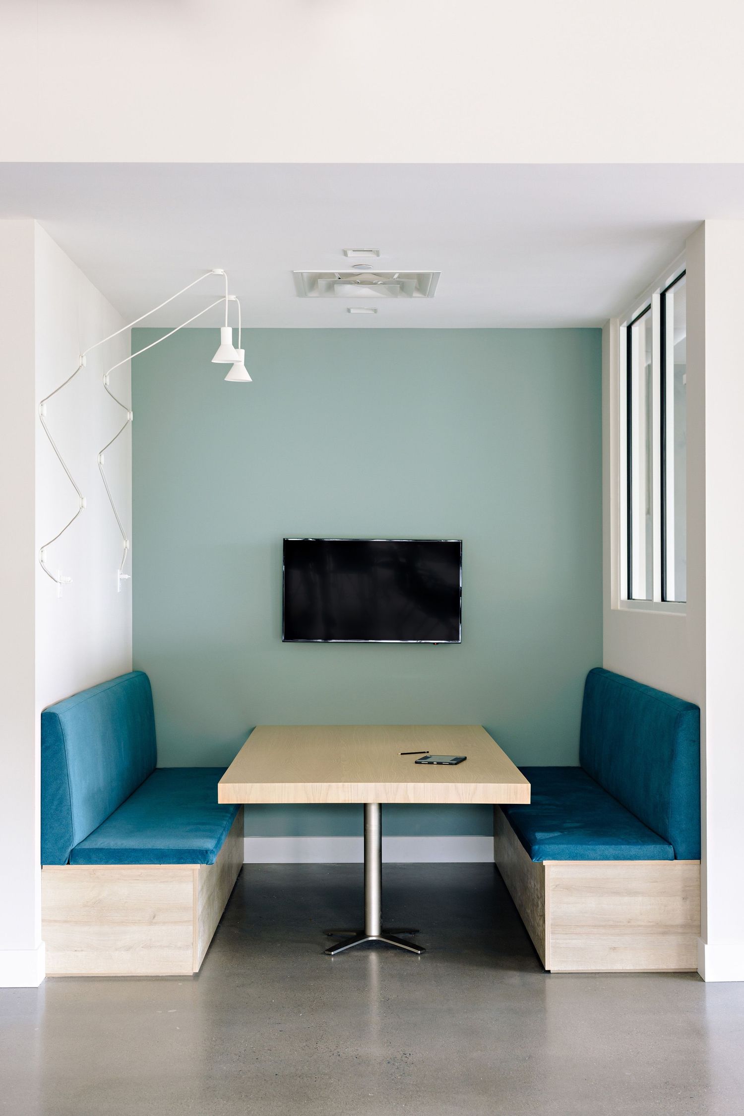 Modern office booth with teal upholstered benches, light wood table, and wall-mounted TV against a mint green accent wall.