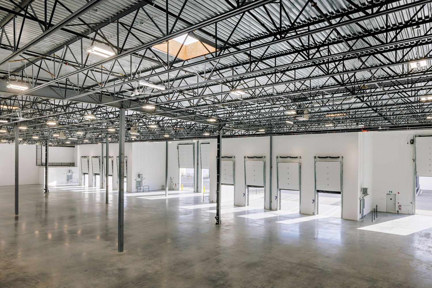 Large empty industrial warehouse interior with exposed ceiling structure, concrete floors and white walls with loading bays.