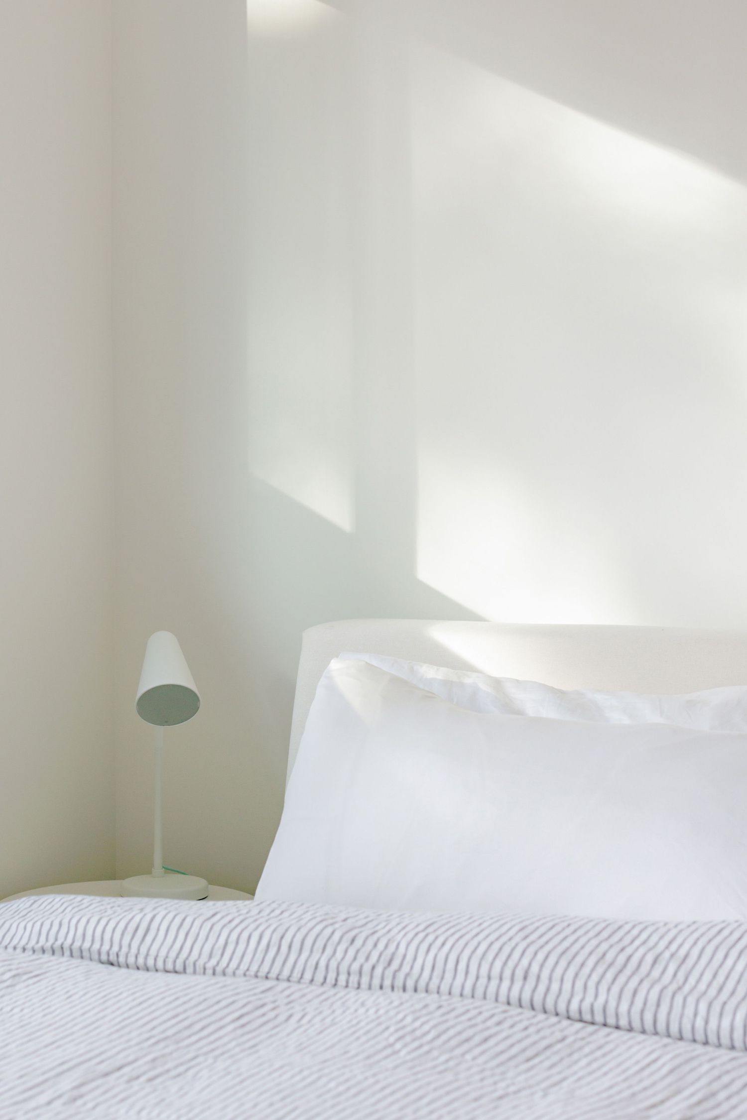 Clean white minimalist bedroom interior with crisp bedding, striped duvet cover and modern bedside lamp in natural light.