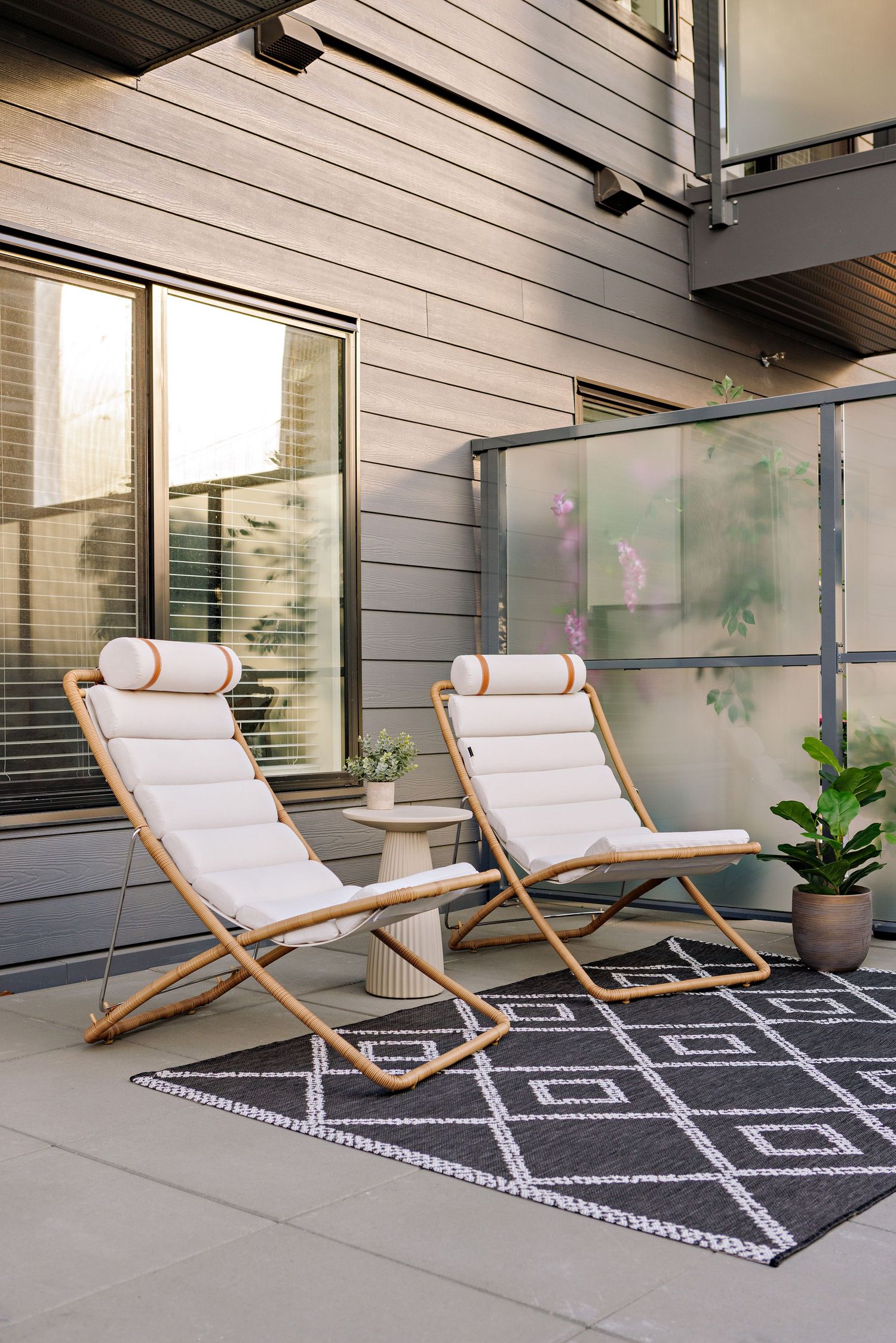Modern patio space with two wooden loungers with white cushions on a geometric patterned rug beside a glass wall.