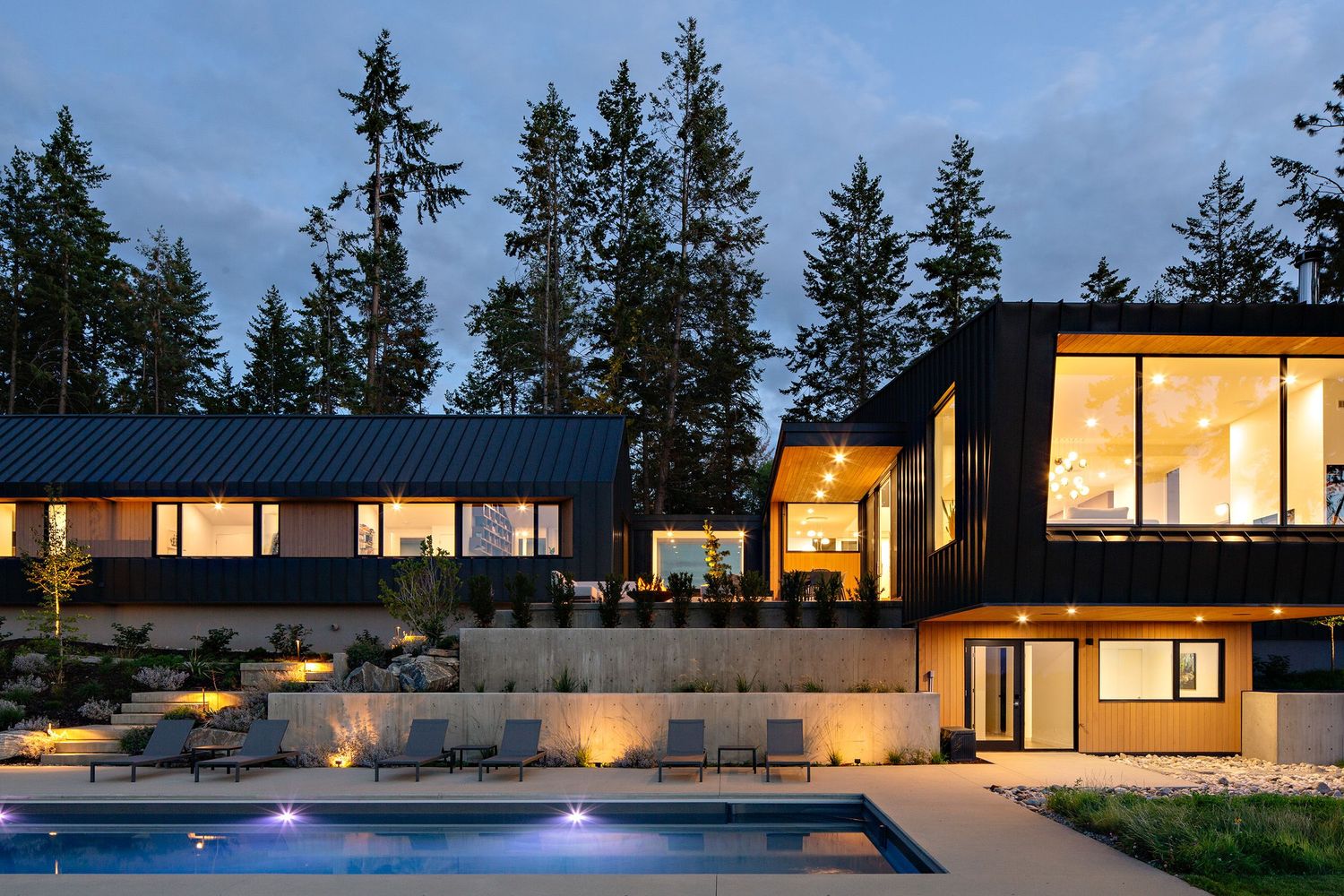 Contemporary luxury home with illuminated windows and swimming pool at dusk against evergreen forest backdrop.